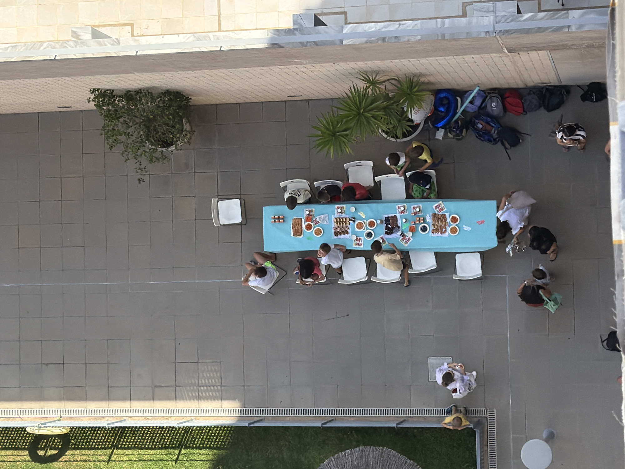 This is an aerial shot of a gathering of people at an outdoor event, possibly a party or meal. A group of about ten people are seated around a long, blue-clothed table laden with food and drinks. Several individuals are standing and mingling. The scene appears to be on a paved patio area, with a building or wall in the background. There are also potted plants and a patch of green grass with some lounge chairs visible near a body of water, suggesting a recreational area. Several backpacks and a large blue balloon are also present. The lighting suggests it is daytime, with shadows cast on the ground, indicating it might be late morning or afternoon. The people's postures do not convey strong emotions.