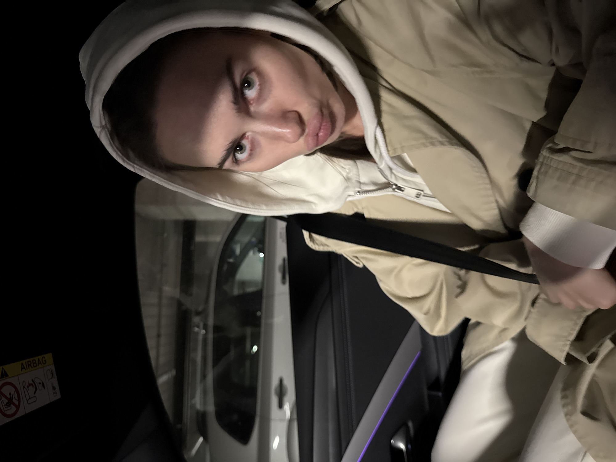 A woman wearing a beige hoodie and jacket is seated inside a vehicle. She is looking at the camera with a pursed-lip expression. She is wearing a seatbelt. The interior of the car is black with a white door panel visible. An airbag warning sticker is visible on the vehicle's interior. The location is not discernible from the image.