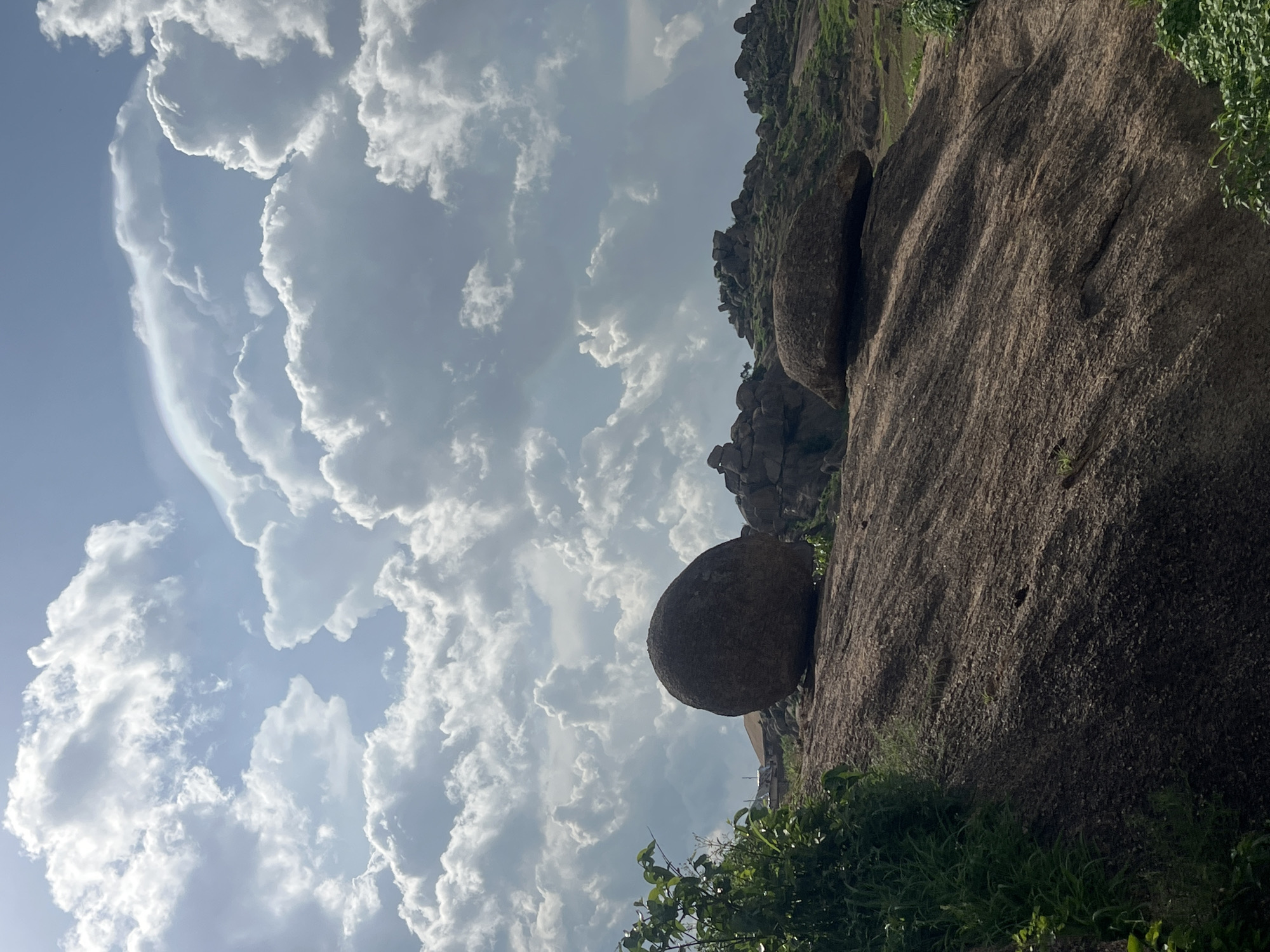 The media file captures a dramatic landscape in Jos, Nigeria, characterized by a rugged, rocky terrain against a backdrop of a dynamic sky. The primary subjects are large, weathered boulders and rock formations, with one particularly prominent, spherical boulder perched precariously on a slope. The rocks appear to be ancient, with textured surfaces suggesting erosion over time. Lush green vegetation, including grass and some leafy bushes, grows sparsely around the base of the rocks and on the hillsides, adding a touch of vibrant life to the scene.
The sky is filled with dramatic, voluminous clouds. These clouds are illuminated by sunlight, creating a bright, almost ethereal effect, particularly on the left side of the frame where they are dense and billowy, casting some shadows on the rocks. The lighting suggests it is daytime, possibly late afternoon given the angle of the sun and the shadows. The weather appears to be somewhat unsettled, with the dramatic cloud formations hinting at potential rain or a transition in the weather.
The overall mood conveyed is one of natural grandeur and resilience. The immense scale of the rocks and the dramatic sky evoke a sense of awe and perhaps isolation. The image does not contain any people or visible text. The specific location cues of Jos, Nigeria, combined with the characteristic rock formations and vegetation, help to ground the scene in a particular geographical context. The composition, with the rocks dominating the right side and the sky filling the left, creates a striking visual contrast between the solid, earthly elements and the ephemeral nature of the clouds.