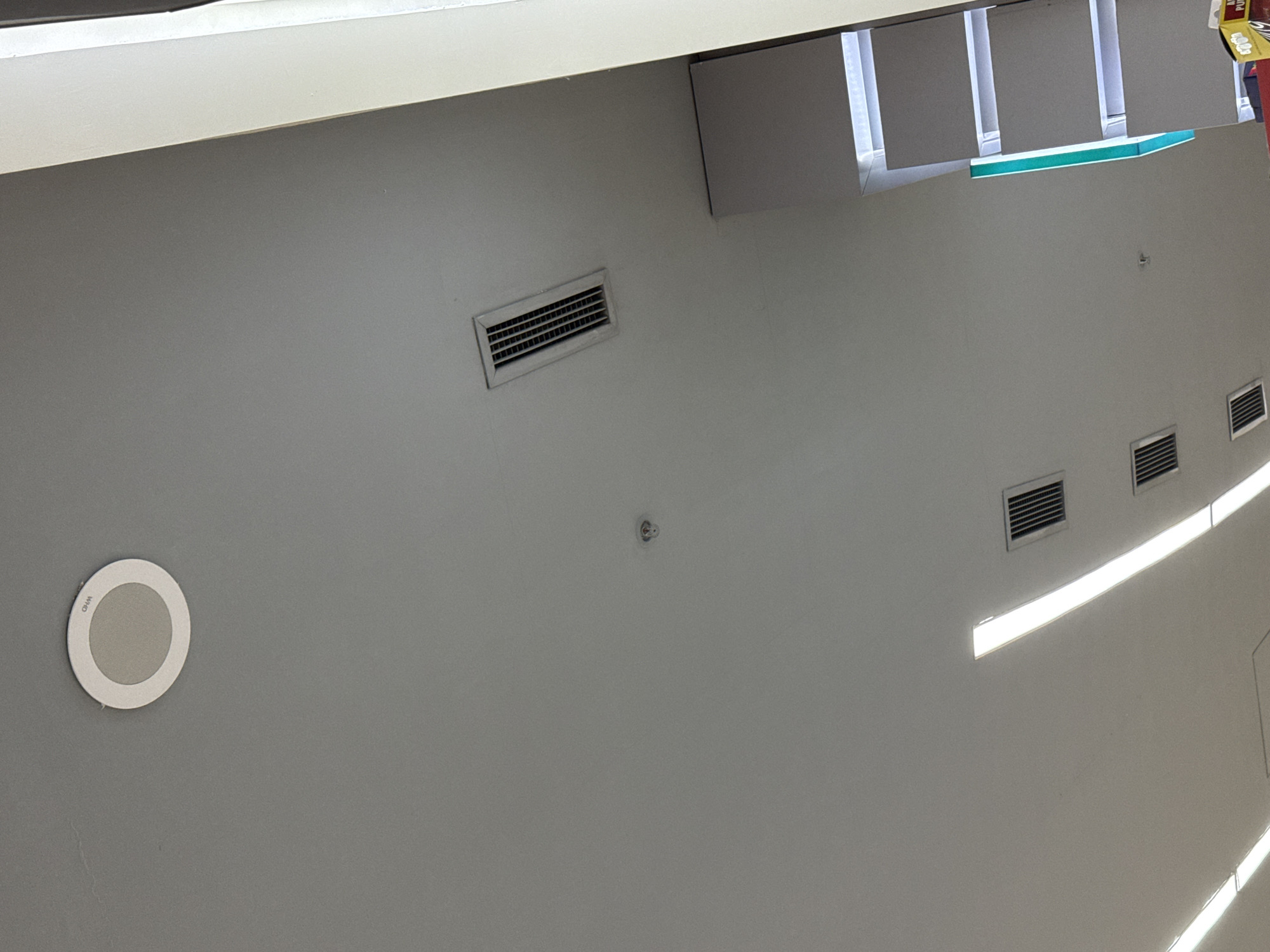 The image displays a modern interior ceiling, photographed from an angled perspective that makes it appear like a wall. The ceiling is predominantly a uniform light grey color. Several architectural and functional elements are visible across its surface.
Three rectangular air vents or diffusers are present, with one in the upper-left area and two aligned horizontally towards the right side of the image. A circular recessed fixture, likely a speaker or a light, is positioned in the lower-left section, bearing the faint text "WXO" on its surface. Near the center of the image, a small, subtle embedded fixture, possibly a sprinkler head or a smoke detector, is observable.
Linear light fixtures, emitting bright white illumination, run across the upper and middle-right portions of the ceiling, providing artificial lighting to the space. The top edge of the overall image reveals the underside of an overhead structure or bulkhead, which has several square-shaped elements and an additional light fixture with a prominent green accent strip. A small portion of colorful commercial packaging is just visible at the extreme top-right corner, hinting at a retail or public building environment. No people or specific activities are depicted in the scene.
