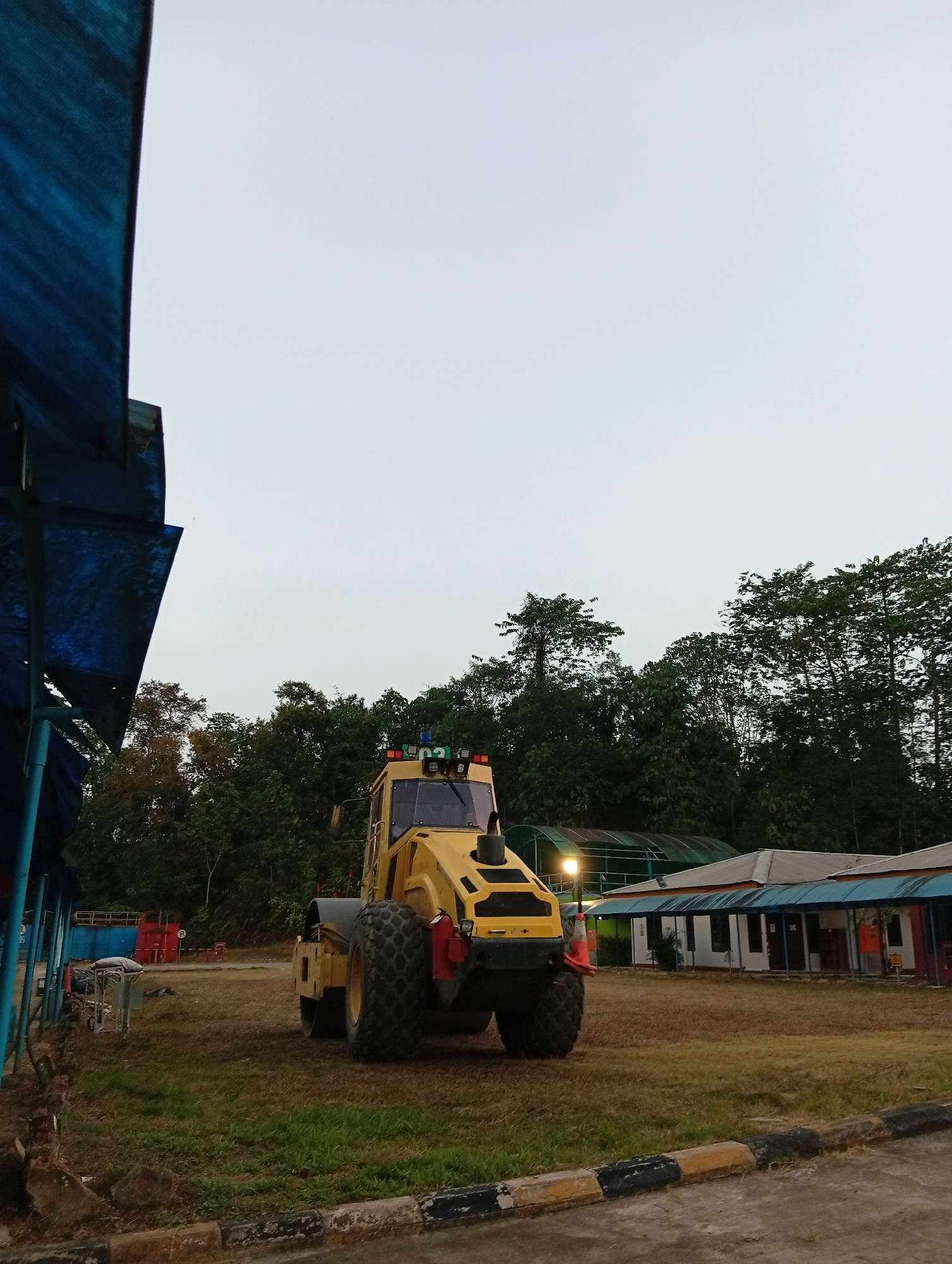 A yellow steamroller is parked in a grassy field. The steamroller is large and has two large wheels. It is facing the camera and is in the center of the image. In the background, there are trees and a building. The sky is overcast.
