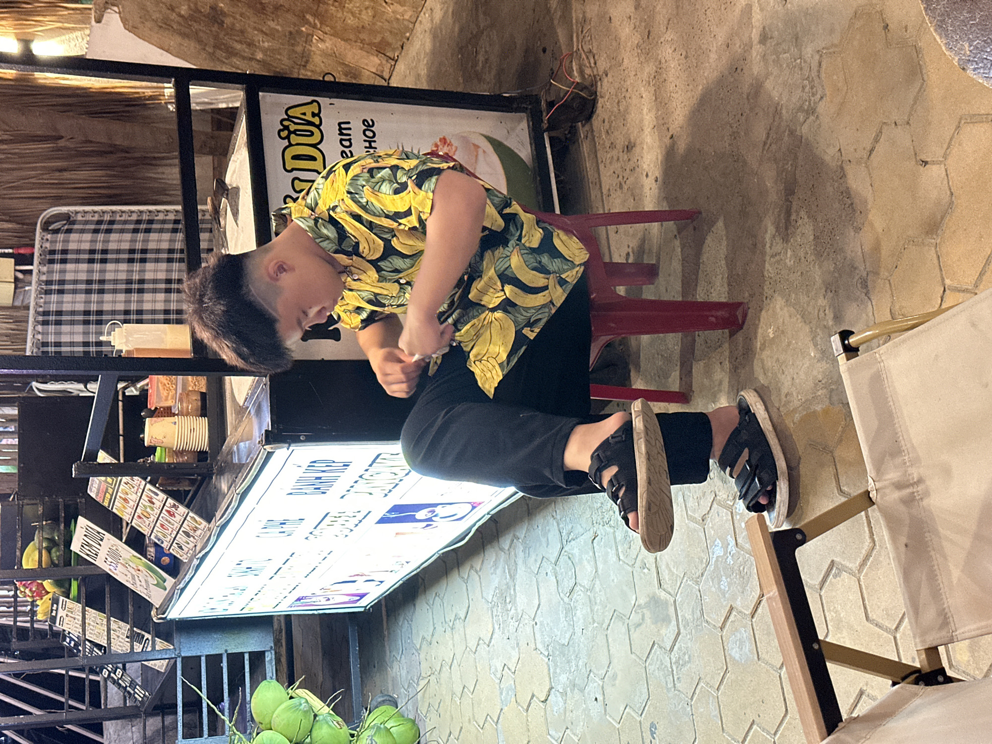 In Phu Thuy Ward, Vietnam, a young male with short, dark, styled hair is seated on a red plastic stool at an outdoor stall. He is wearing a vibrant yellow shirt adorned with a banana print and black pants. He is looking down and appears to be engaged in an action with his hands, possibly adjusting something. His feet are clad in black sandals. In the background, there is a food stall with illuminated signage displaying Vietnamese text and images of food and drinks. Coconuts are visible to the left of the stall, and a checkered cloth is draped in the background. The ground is paved with hexagonal tiles.