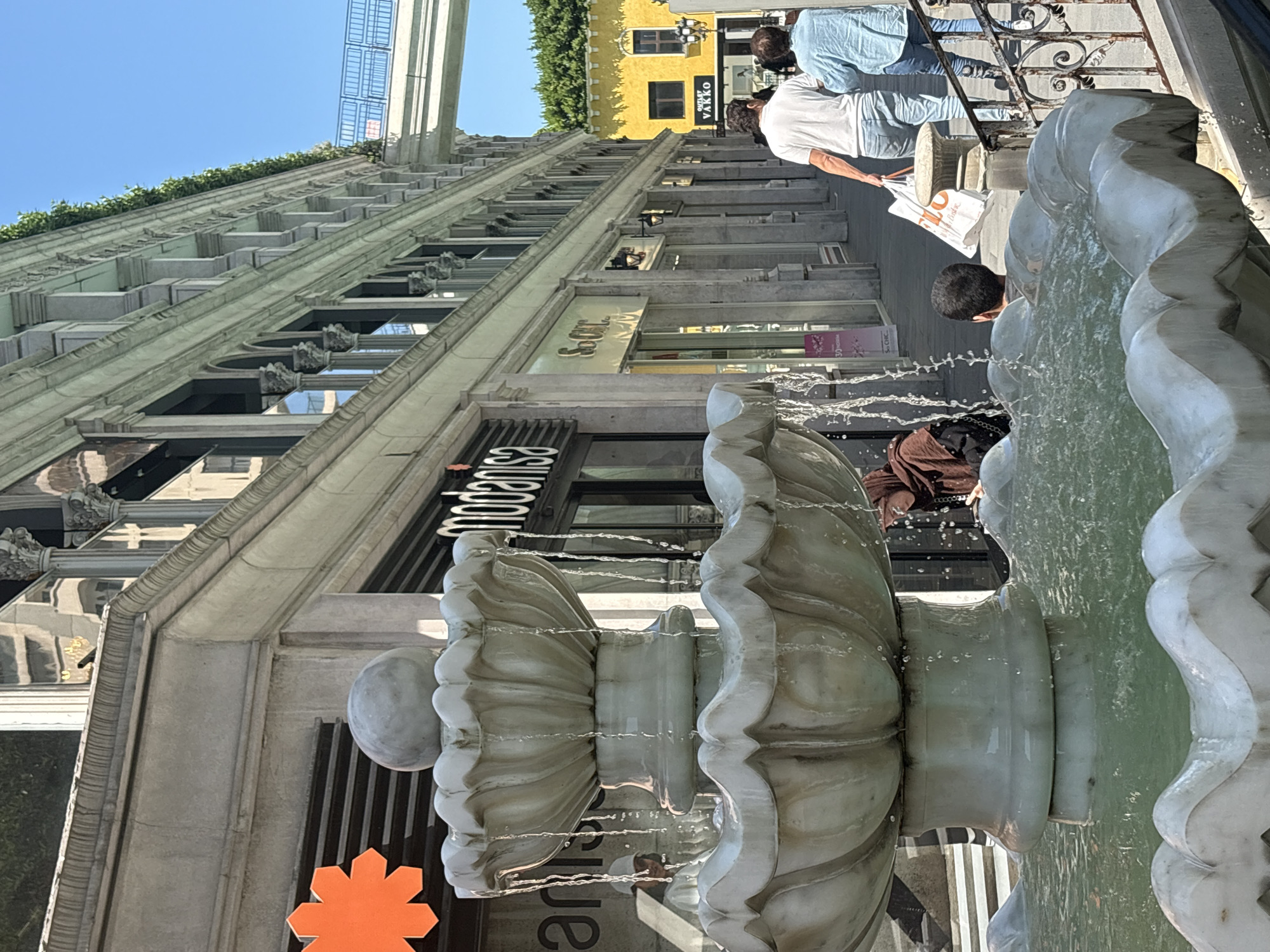 This image captures an outdoor commercial street scene in Gaziosmanpaşa, Turkey, under clear daylight.
In the foreground, a multi-tiered, white marble fountain dominates the lower left, with water actively flowing from its upper basins into the large, greenish-water-filled lower basin. A spherical finial tops the uppermost tier.
The midground features a paved pedestrian area lined with shops. To the left of the fountain, partially visible behind it, a reflection of a person in a white cap appears in a shop window. Further left, an orange, star-shaped logo is partially visible. The shop directly behind the fountain is identified by a prominent black sign displaying "modanisa". A woman in a brown hijab and dark clothing is partially visible near the fountain's left side, looking downward. To the right of the fountain, a young man with dark hair is seen from behind, looking into the fountain's basin. A black metal railing separates the fountain area from the adjacent pedestrian path. On the path, two individuals walk away from the viewer: a man in a white t-shirt and blue jeans, carrying a white shopping bag with an orange logo, is followed by a woman in a light blue button-up shirt and blue jeans. Further along the path, another shop entrance marked "S.O. Chic" is visible.
The background consists of a long, multi-story building running parallel to the street on the left, characterized by numerous classical columns, arched windows, and decorative cornices. Green foliage lines the top edge of this building. A light-colored pedestrian bridge with blue railings spans the upper right portion of the image, connecting to another structure. Beyond the shops, a distinctive yellow building with a dark roof and multiple windows is visible, featuring a black sign that reads "Outlet VAKKO". The sky above is a clear, bright blue.