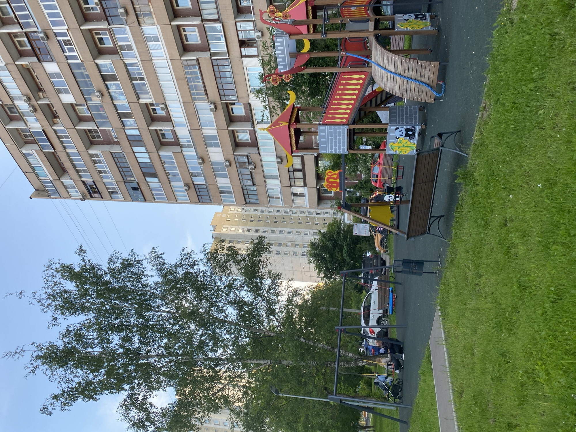 This image captures a vibrant residential scene, likely in Khimki, Russia, focusing on a modern playground set against a backdrop of multi-story apartment buildings. The time of day appears to be late afternoon or early evening on a clear, sunny day, indicated by the lighting and the blue sky.
In the foreground, a colorful playground takes center stage, featuring a multi-level wooden structure with bright red and yellow accents. It includes slides, climbing elements, and a bridge with a blue rope. Several grey panels on the play equipment display stylized black and white panda faces. Benches are integrated into the playground design, which sits on a dark rubberized surface. Adjacent to the playground, there are metal swings, and a person dressed in dark clothing is seen sitting on a bench near them, attending to a baby in a stroller.
The background is dominated by a large, light brown apartment building with numerous windows and several air conditioning units visible on its facade. Another, lighter-colored apartment building stands further in the distance. A tall, leafy tree, possibly a birch, occupies the left side of the frame, its green foliage contrasting with the urban structures. Power lines are faintly visible against the sky.
Cars are parked in a paved area beyond the playground, including a white car and a dark-colored vehicle. Green grassy areas frame the playground and pathways, adding a natural element to the urban environment. The scene depicts a typical residential area, bustling with daily life and providing recreational space for children. No clearly readable text is discernible on signs or objects within the image, apart from abstract graphics on the playground.
