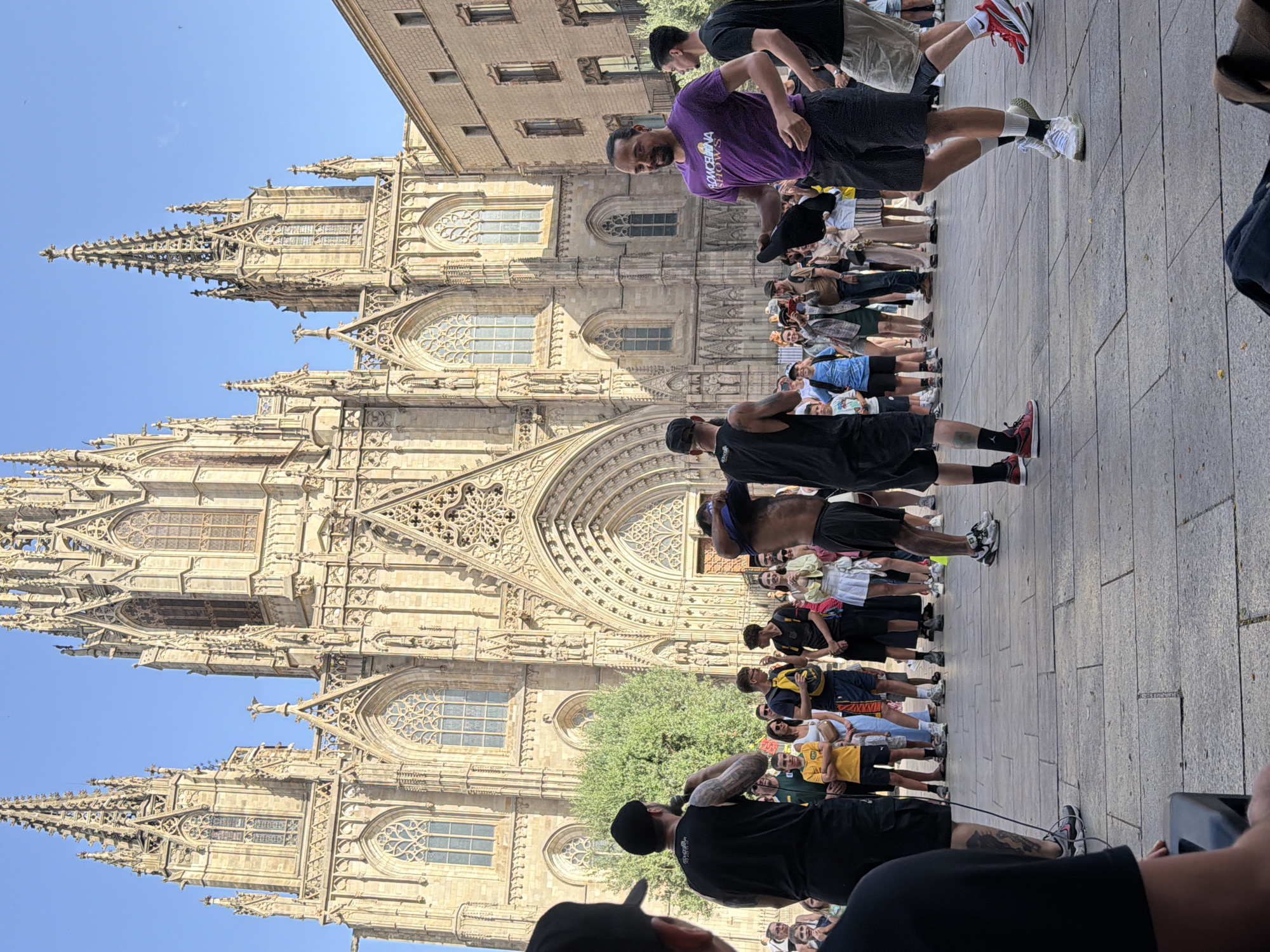 In a vibrant outdoor scene in Barcelona, Spain, a group of people are gathered in front of an impressive, ornate Gothic cathedral. The building's facade features intricate stonework, tall spires, arched windows, and detailed sculptures, suggesting it is a historical landmark. The sky above is a clear, bright blue, indicating a sunny day, likely midday given the strong shadows cast on the ground.
The people in the foreground and middle ground appear to be participating in or watching an event. Some individuals are dressed in casual, athletic wear, with several men in t-shirts and shorts, suggesting a performance or active gathering. One man in the mid-ground wears a bright purple t-shirt with visible white text that reads "BARCELONA SHOWS". Another individual, in the far foreground, is wearing a black t-shirt and a black cap, and appears to be speaking into a microphone, possibly as an announcer or performer.
The ground is paved with large, square stones, typical of European city plazas. A line of people stands facing the cathedral, some with hands clasped behind their backs, others looking towards the possible performance. The atmosphere seems engaged and lively, with a mix of participants and spectators. The overall impression is one of a cultural event or street performance taking place in a historically significant urban setting.