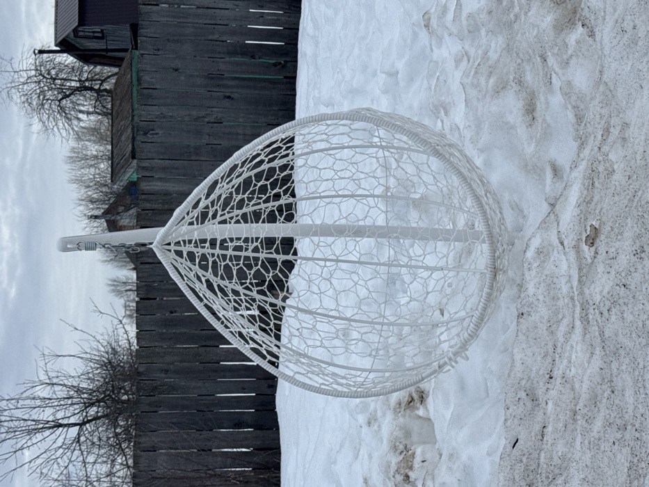 This image displays a white, teardrop-shaped hanging egg chair positioned outdoors in a snow-covered area in the city of Chaikovsky, Russia. The chair features a white woven lattice design and is suspended from a white metal stand by a visible metal chain and spring. The ground beneath and around the chair is covered in dirty, partially melted snow, revealing patches of dark earth. In the background, a weathered wooden fence runs horizontally across the scene. Beyond the fence, bare deciduous trees are visible against an overcast sky. Several structures are present behind the fence, including a dark-colored building with a sloped roof and a chimney on the right, and a smaller, light green-blue wooden structure further to the left. No individuals or active subjects are present in the scene.