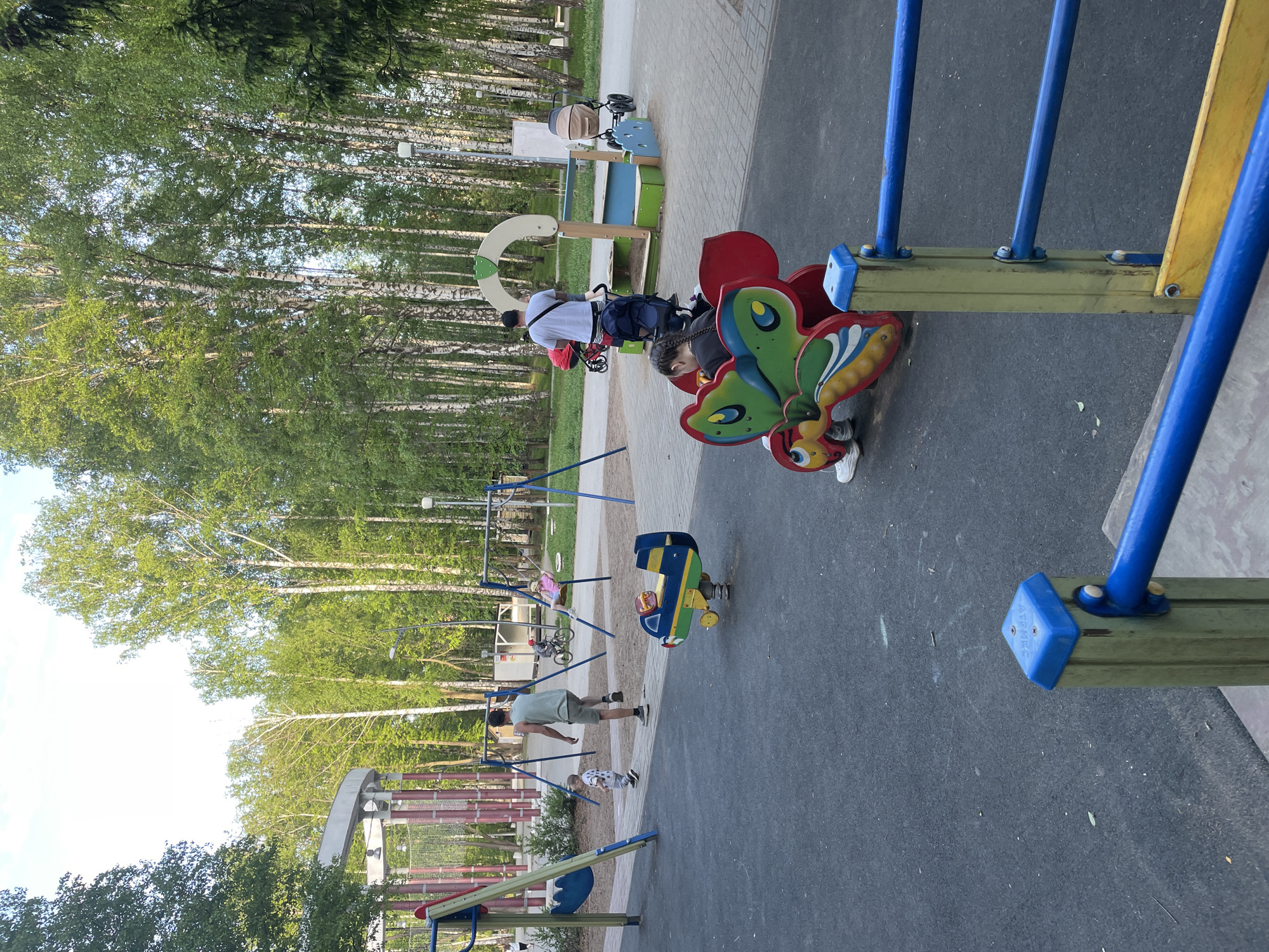 The image depicts a playground in the city of Perm, Russia. A man and a child are on a butterfly-shaped ride. A man is helping a child on the swings. There are trees in the background.
