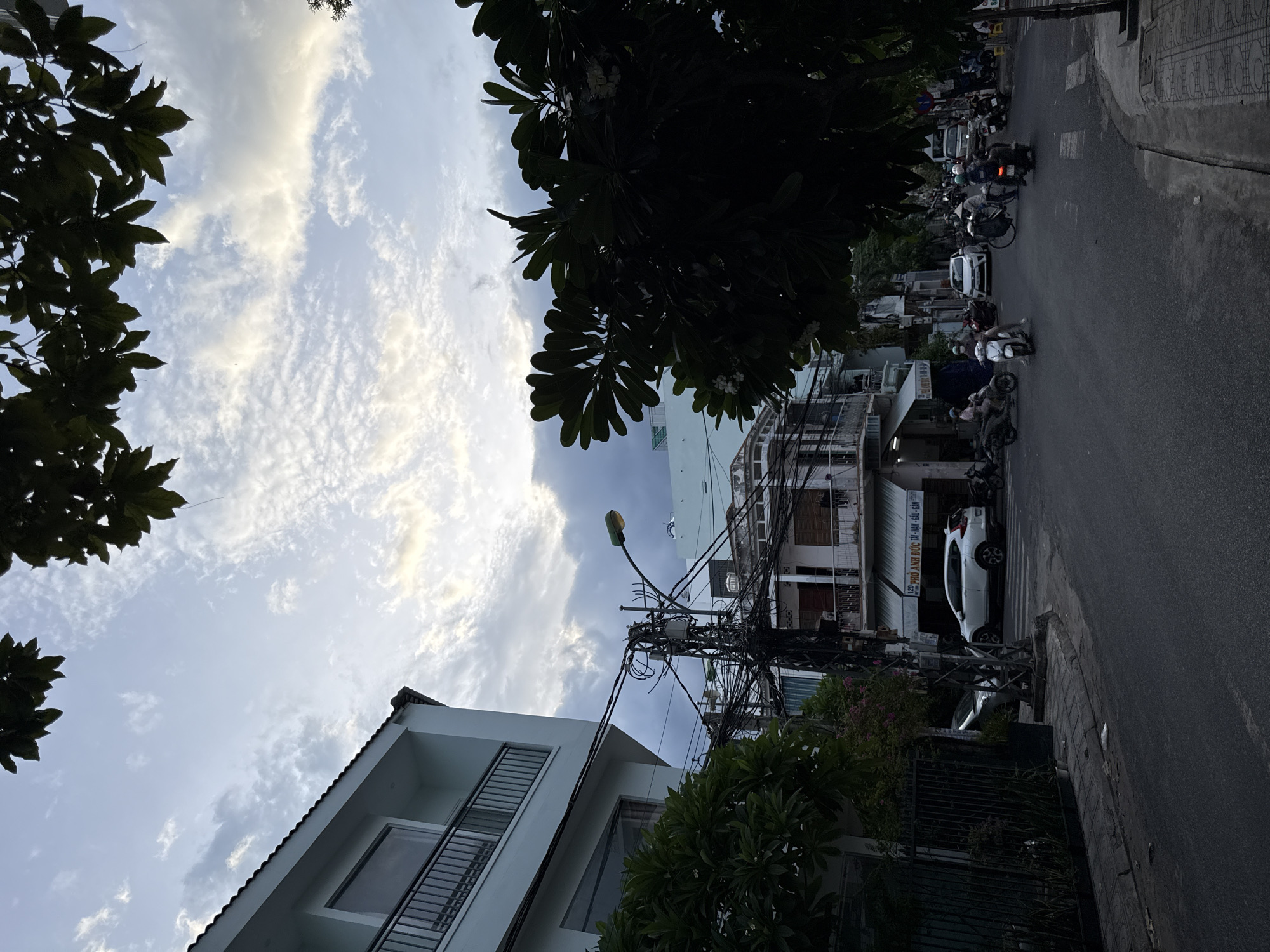 This image captures a street scene in Nha Trang, Vietnam, under a partly cloudy sky. On the left, a modern, light-colored building with balconies and dark railings stands prominently. Adjacent to it, a dense tree with dark foliage partially obscures the view of other buildings. A complex network of electrical wires is suspended from a utility pole, adding a sense of urban infrastructure to the scene.
The street is paved and appears to be moderately busy with traffic. Several motorcycles, some with riders, are visible, suggesting the common mode of transportation in the area. One white SUV is parked or driving on the street. Further down the road, more buildings, commercial storefronts with awnings, and a variety of motorcycles are present. The architectural style of the buildings suggests a blend of modern and older constructions. The overall impression is that of a typical day in a bustling Vietnamese city.