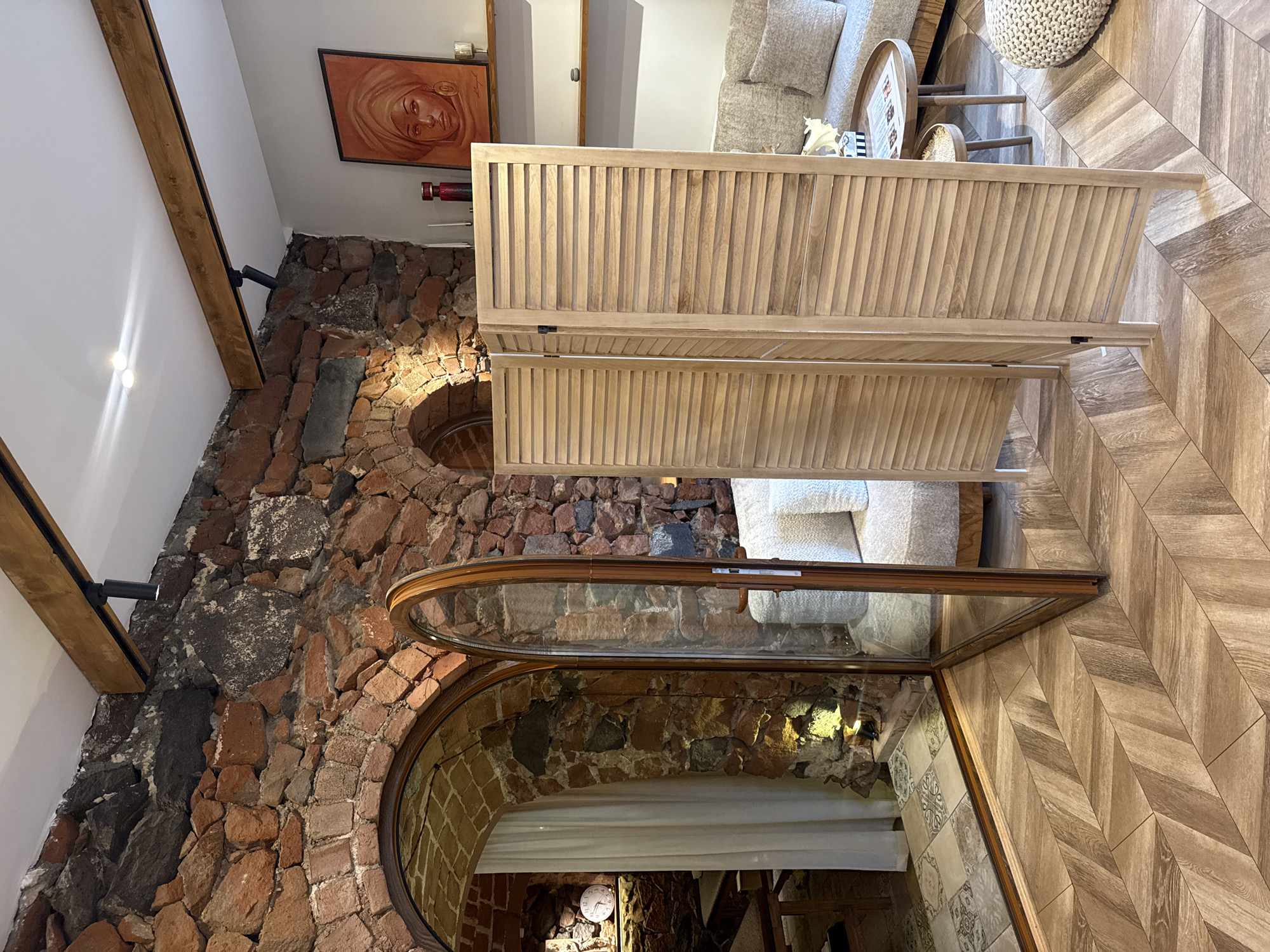 The image captures an interior space in Yerevan, Armenia, characterized by a blend of rustic and modern design elements. The dominant features include a rough-hewn brick wall with arched openings, one of which contains a framed glass partition. A light-colored wooden screen stands in front of the brick wall. The floor is covered in light brown herringbone-patterned tiles. Furniture such as a light-colored couch and a small round table are visible, indicating a living or lounge area. An artwork of a woman's face hangs on the wall. Lighting fixtures are attached to wooden beams in the ceiling, suggesting an intentional design for the space's ambiance.