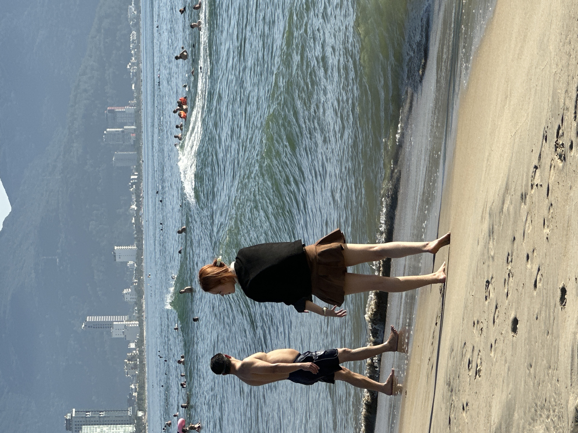 An eye-level, outdoor, vertical shot captures a wide view of My Khe Beach in Da Nang, Vietnam, on a sunny afternoon. The foreground is dominated by a sandy beach with footprints, leading to the ocean. Two people stand near the waterline. On the left, a bare-chested man with dark hair and a headband faces away from the camera, wearing dark blue swim trunks. On the right, a young woman with reddish hair tied back, facing the man, wears a black long-sleeved top and a brown skirt. She has her left hand raised. The ocean is a vibrant blue-green, with moderate waves breaking near the shore. Numerous people are visible swimming and playing in the water further out. In the distance, the city skyline of Da Nang rises, featuring tall buildings and smaller structures, backed by lush green mountains under a clear blue sky. The overall scene conveys a sense of leisure and activity on a popular beach.