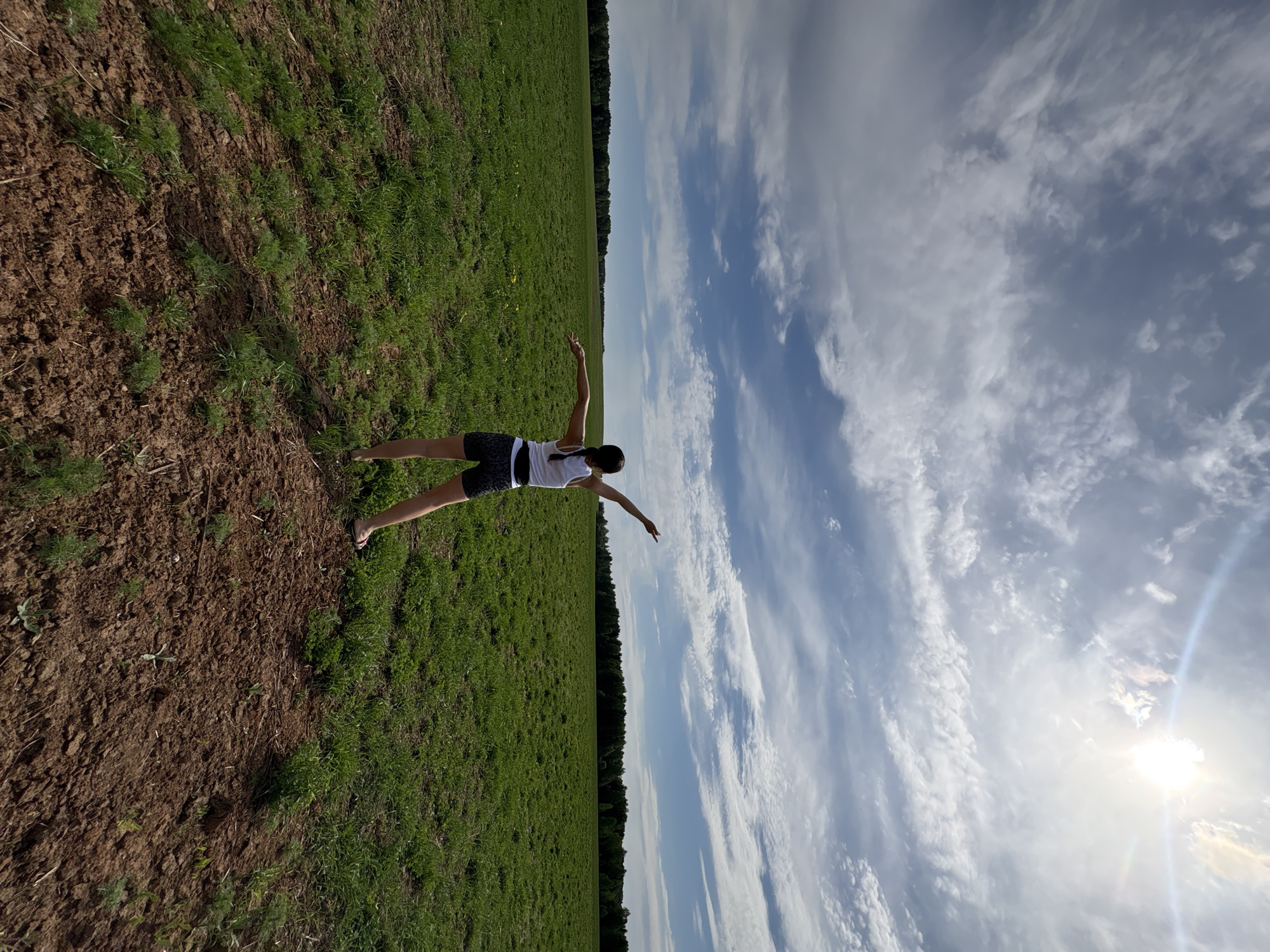 A woman stands in a field under a partly cloudy sky, with her arms outstretched. The field is divided into two sections: one with green grass and the other with exposed dirt. A line of trees marks the horizon. The sun shines brightly in the sky. The location is Чумна, Russia.