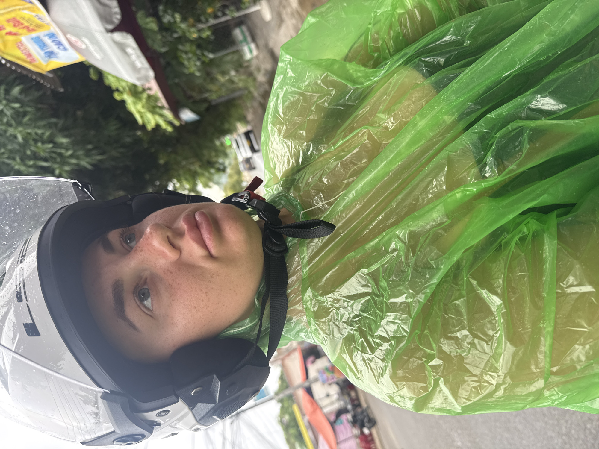 A woman is riding a motorcycle wearing a white helmet and a bright green poncho. She is looking upwards and to the right, with her face partially obscured by the helmet. The background shows a street scene in Nha Trang, Vietnam, with buildings, trees, and other vehicles visible. The weather appears to be overcast or rainy, as indicated by the wet surfaces and the woman's poncho.
