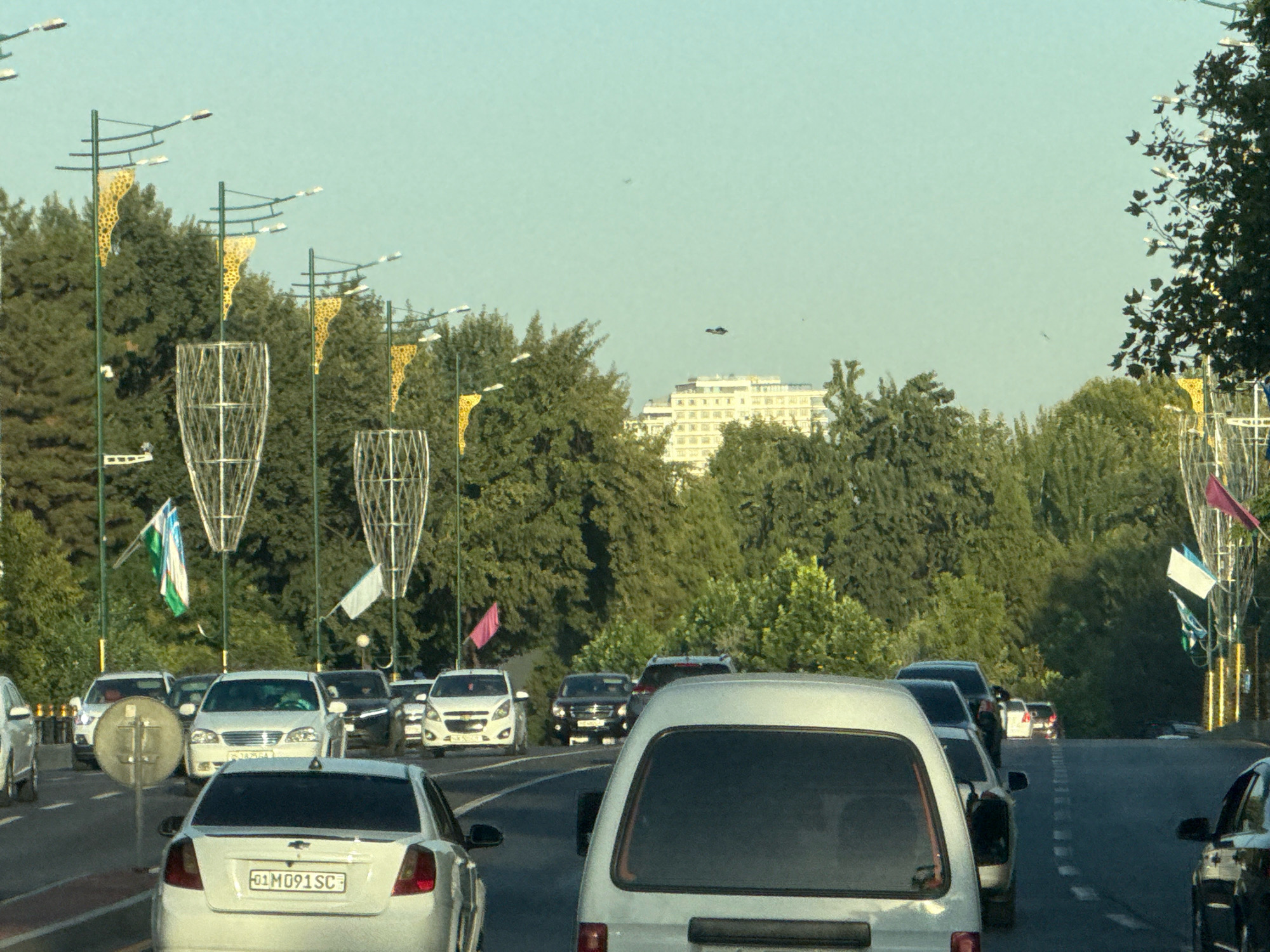 The image depicts a street scene in Tashkent, Uzbekistan, likely during daylight hours. In the foreground, a white van is traveling in traffic. Behind it, several cars are visible, including a white sedan with a license plate starting with "01 M091 SC", suggesting it is registered in Tashkent. The road is lined with trees and decorative street lamps with yellow, cone-shaped elements. In the background, a tall, white building with multiple floors is visible, and flags are flying from poles along the street. There are also some darker objects in the sky, possibly birds or possibly distant aircraft. The overall atmosphere is that of a typical urban environment with moderate traffic.
