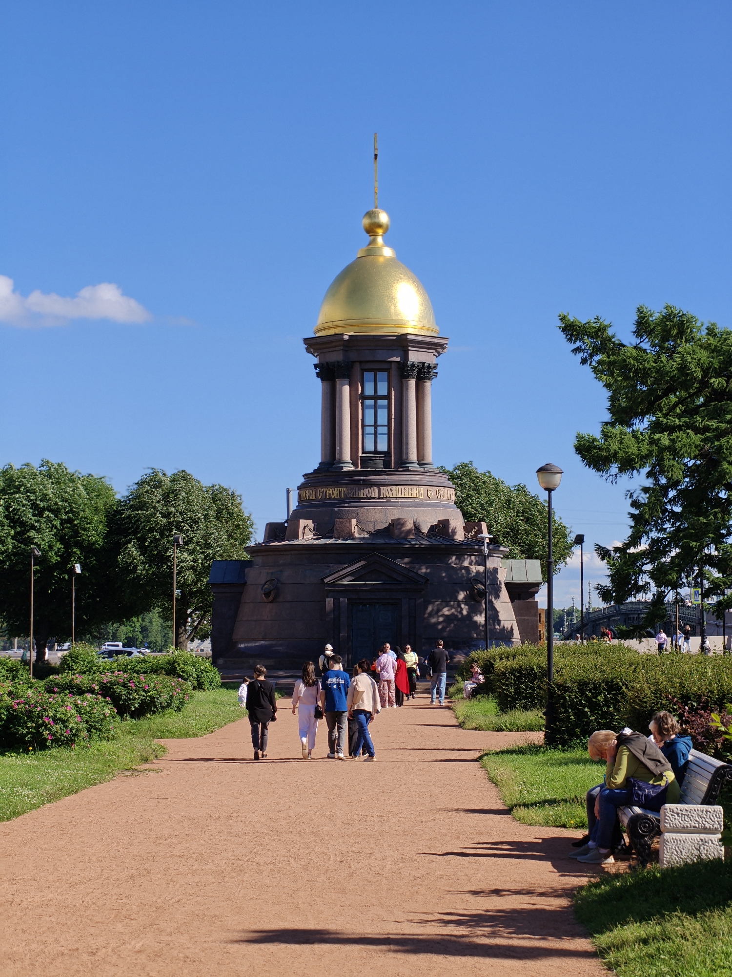 This is a daytime, vertical, medium shot of the Chapel of the Virgin Mary, also known as the Church of the Intercession of the Virgin Mary, in St. Petersburg, Russia. The chapel is a circular, stone structure with a prominent gold dome topped by a spire. It is surrounded by lush green trees and manicured bushes. A gravel path leads towards the chapel, with several people walking on it. On the right side of the path, two people are sitting on a bench. The sky is a clear blue with a few white clouds. The overall atmosphere is peaceful and serene. The inscription on the chapel reads: "СВЯТОЙ ПОКРОВИТЕЛЬНИЦЫ СУДЬБЫ" which translates to "Holy Patroness of Fate".