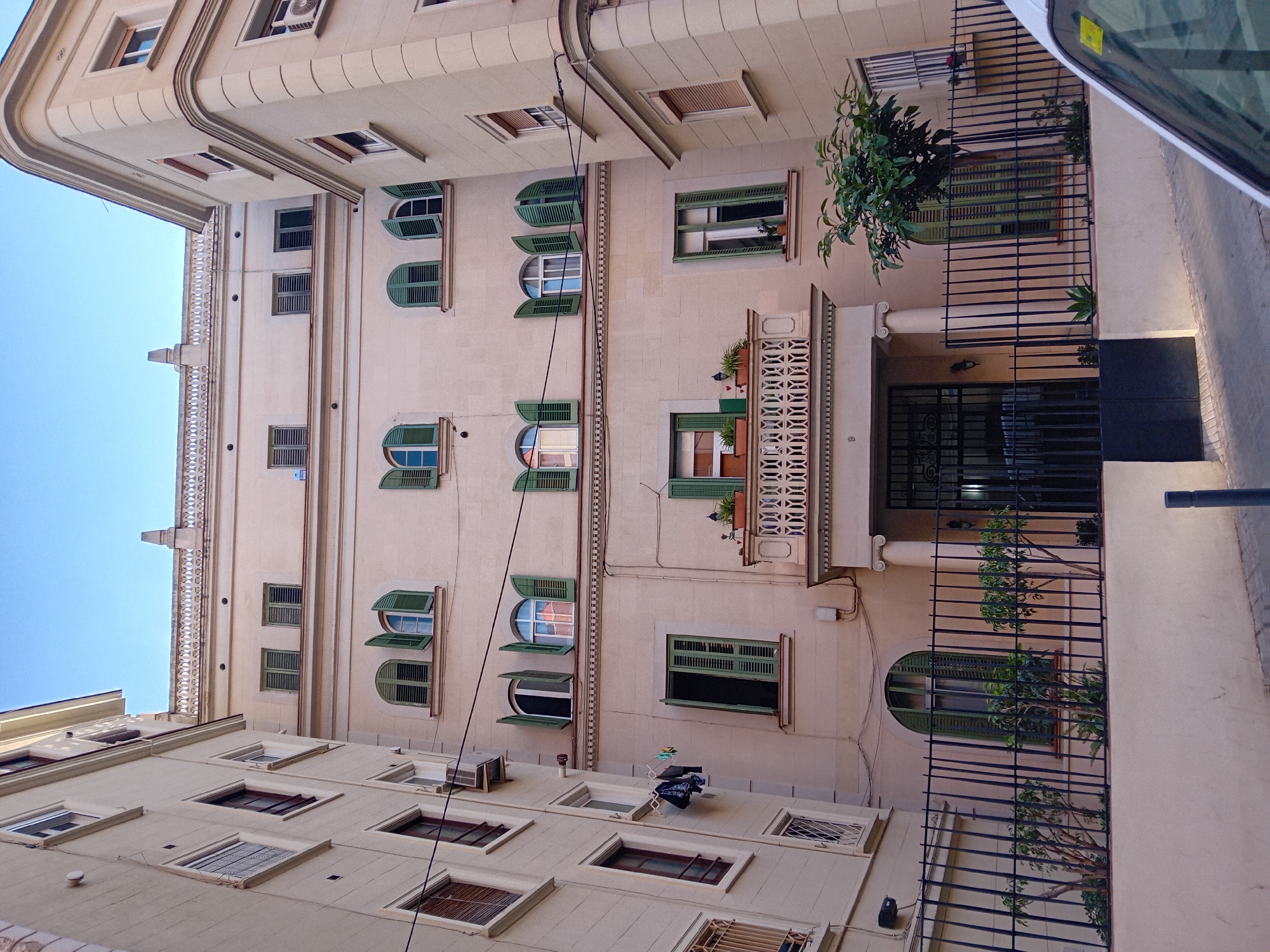 The image captures an upward view of a multi-story residential building facade from a street level in Barcelona, Spain. The building features a light beige or cream-colored exterior with numerous windows and balconies. Many windows are fitted with traditional green wooden shutters, some open, some closed, and vary in shape from rectangular to arched.
Several balconies adorn the facade, some with intricate white patterned railings and potted plants, while others have simpler black metal railings. A prominent balcony with ornate white lattice-work and potted plants is visible in the upper mid-section. Below this, a large entrance gate made of black wrought iron is visible, leading into what appears to be a main entrance. To the right of the gate, part of a white vehicle is visible, with a yellow sticker on its windshield.
Other notable details include a black drainpipe running vertically down the building, air conditioning units mounted near some windows, and a clothes drying rack with laundry hanging on it on a lower-level roof or terrace. A small tree with green foliage is situated near an upper balcony. Architectural details like decorative cornices and balustrades are also visible, particularly on the upper parts of the building. The sky is a clear, light blue, suggesting daylight hours, likely morning or afternoon. The number "8" is faintly visible on a column near the building's entrance. The overall scene depicts a typical, well-maintained urban residential building in a Mediterranean setting.