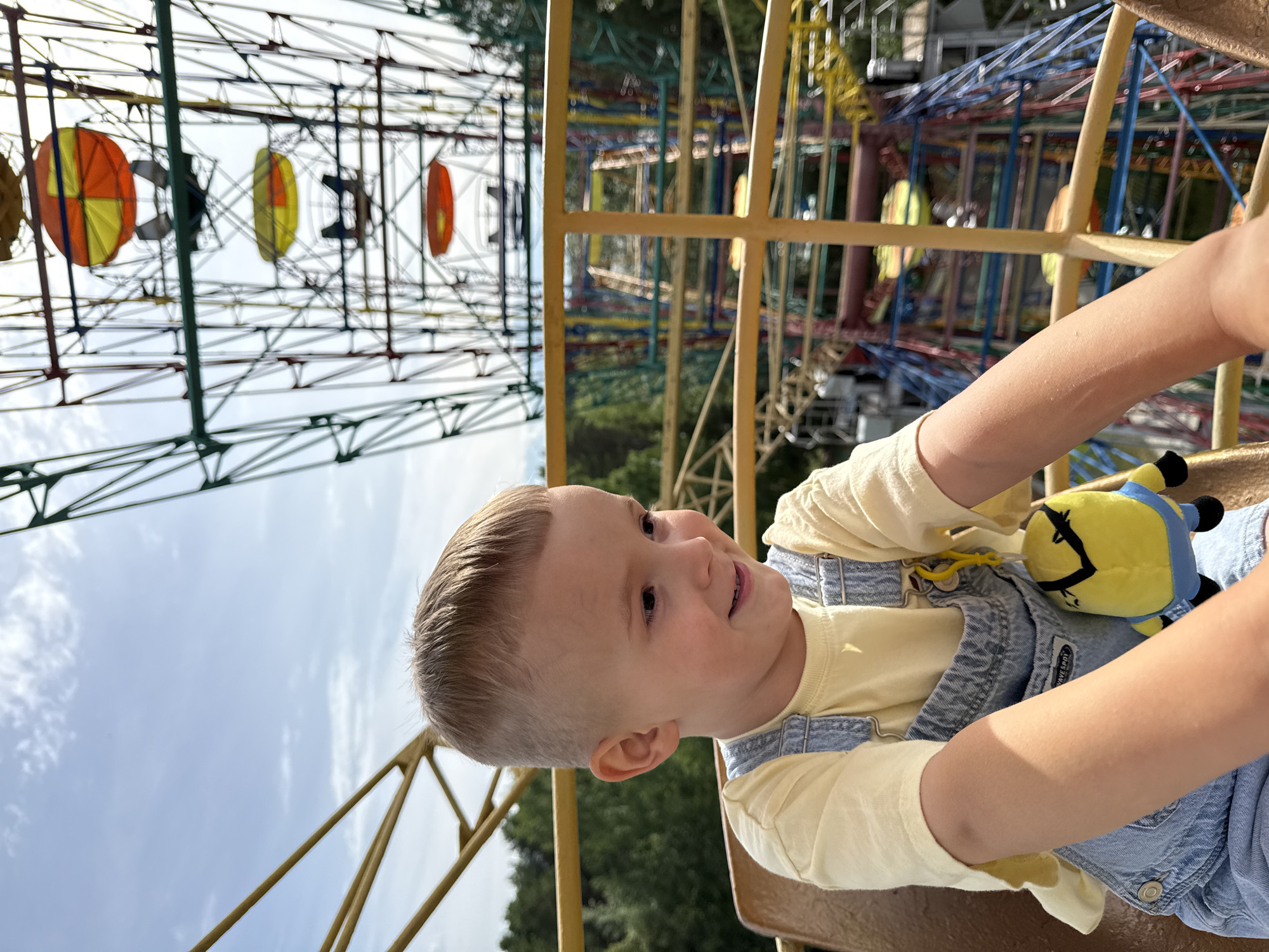 A young boy with blond hair, wearing a yellow t-shirt and denim overalls, smiles as he rides on an amusement park ride. He holds a stuffed Minion toy. In the background, a Ferris wheel with colorful gondolas and a roller coaster with brightly colored tracks are visible. The sky is cloudy. The image appears to be taken during the daytime. The location is identified as Chaikovsky, Russia.
