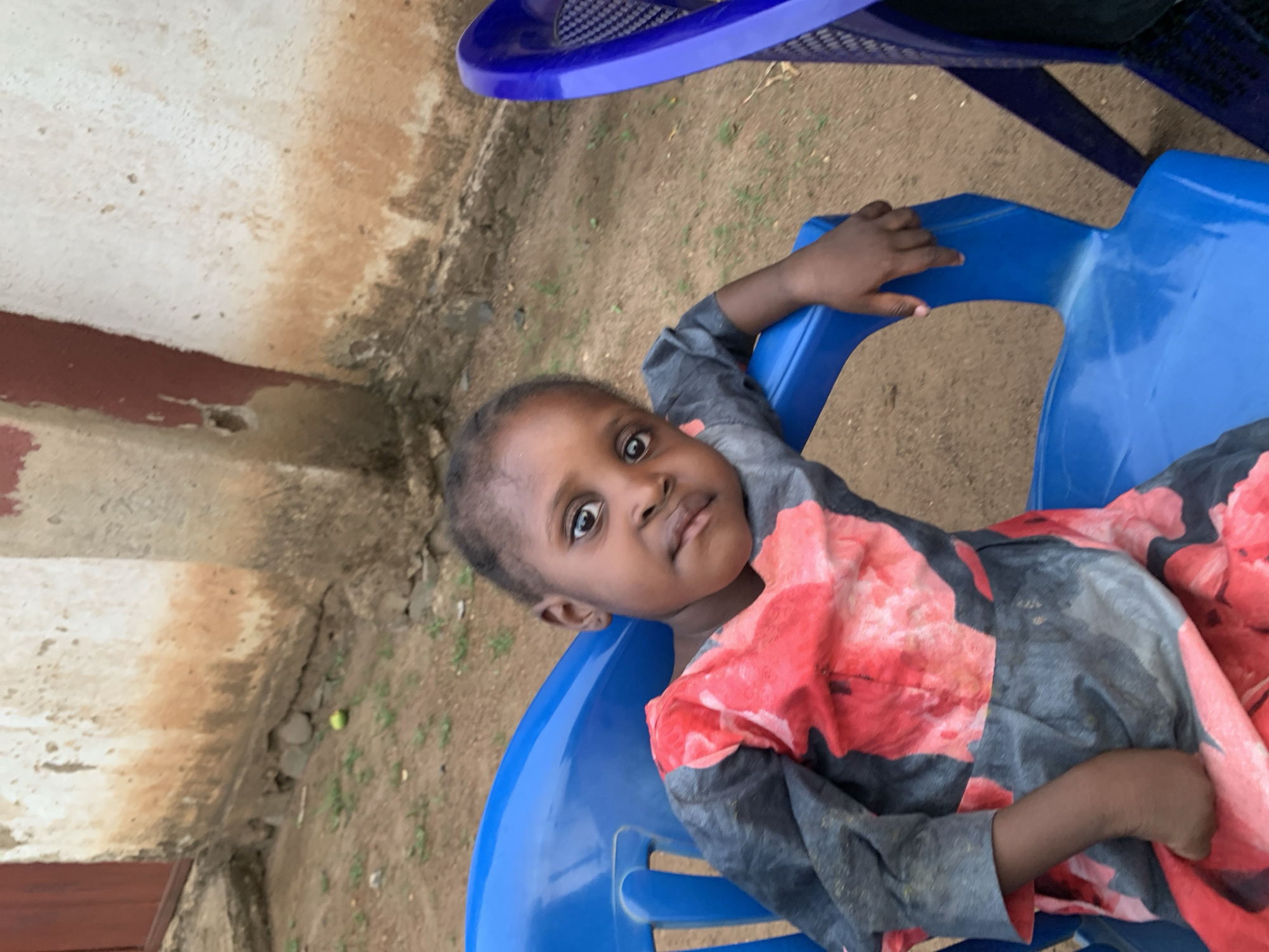 A young child, presumably female, is seated in a bright blue plastic chair. The child is wearing a gray and red floral patterned garment with long sleeves. The child is looking directly at the camera. The setting appears to be outdoors, adjacent to a building with an aged, mottled concrete wall with a red-painted section near the base. The ground is dirt. The image location is Agban, Nigeria.