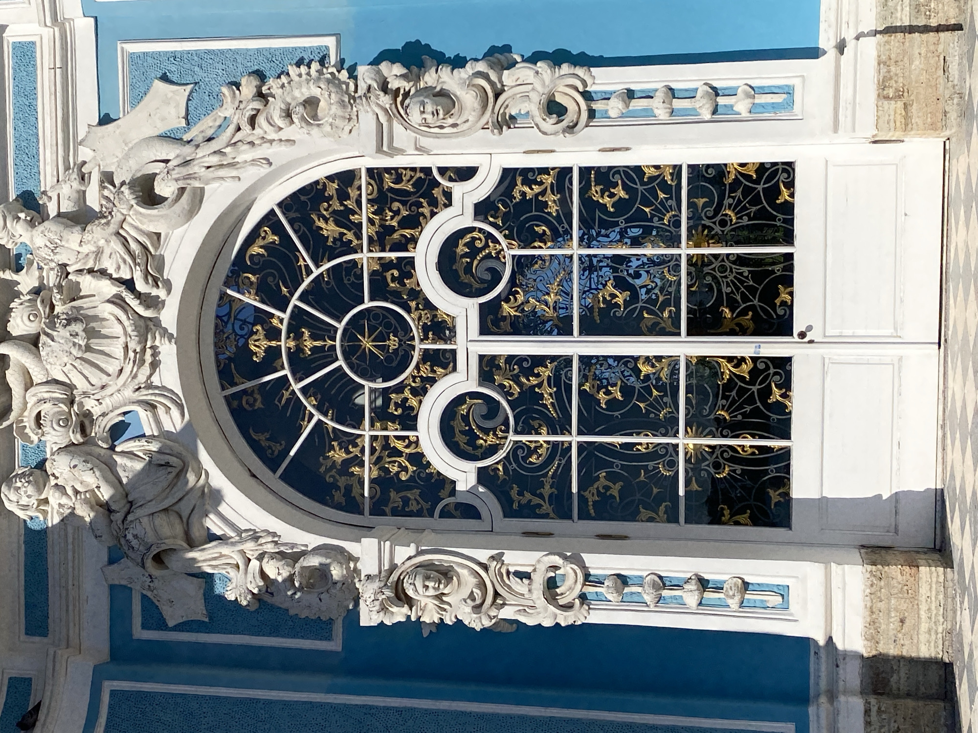 This is a medium shot, eye-level, frontal photograph of a highly ornate, arched doorway with double doors and intricate metalwork, located on the façade of the Catherine Palace in Pushkin, Russia. The building's exterior walls are painted a bright, textured blue with white decorative trim. The doorway itself is framed by elaborate white stucco sculptures. The arch of the doorway is topped with large sculptural elements, including figures of people and mythological creatures. The upper part of the door features an arched window with a white frame and decorative, gold-colored metal grilles featuring swirling floral patterns. The lower part of the door is made of white panels. On either side of the doorway, and integrated into the white frame, are sculpted busts of women. The building's foundation is made of rusticated stone blocks, and the ground in front of the doorway is paved with a checkered pattern of light and dark gray tiles. The sunlight casts a shadow of the architectural elements onto the ground.