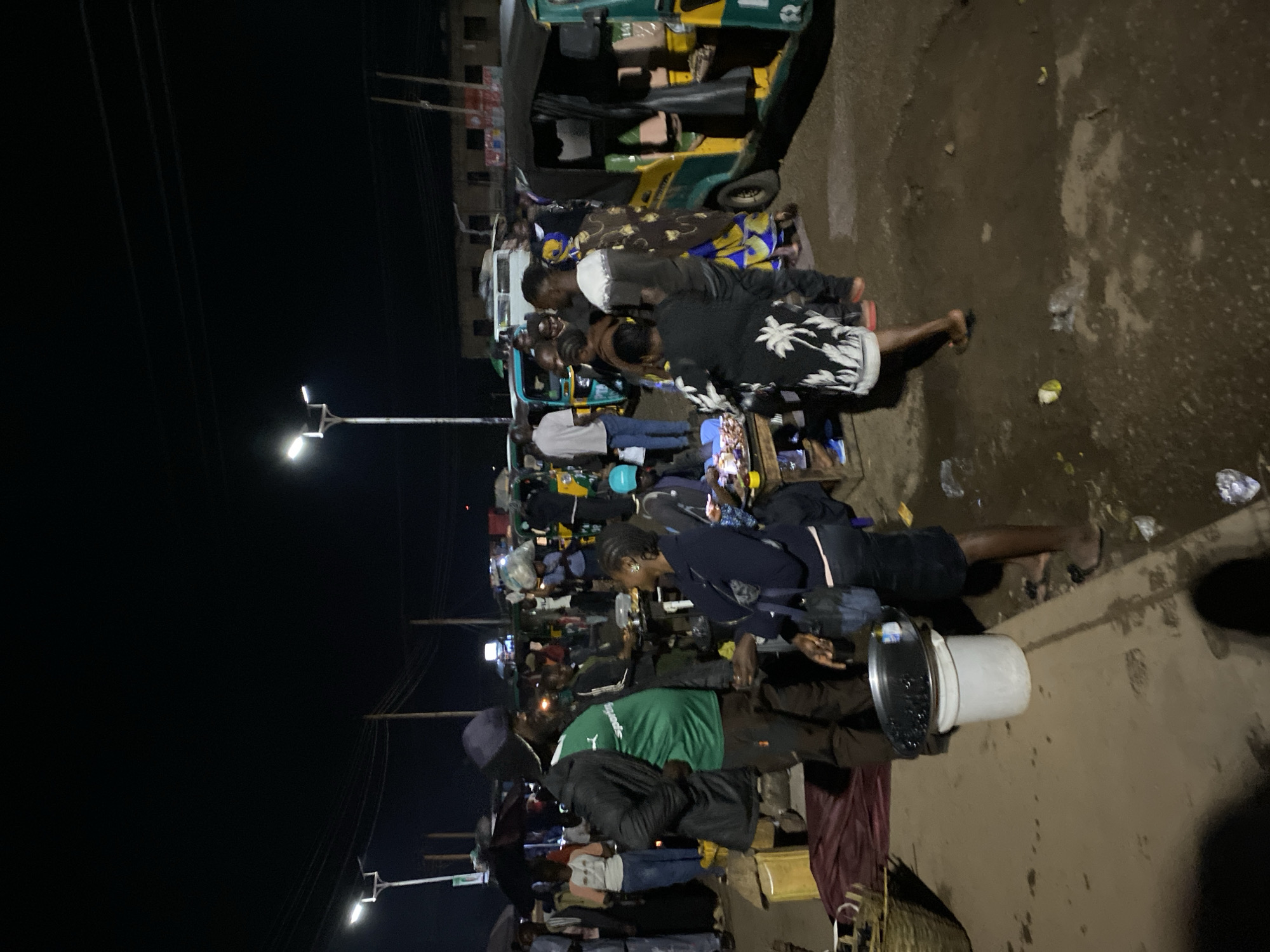 This media file captures a vibrant night scene on an unpaved street in Bukuru, Nigeria. The image, oriented in landscape, depicts a large gathering of people and vehicles under artificial lighting.
**Subjects:** The central focus is a dense crowd of individuals, predominantly men, engaged in various activities. Many are standing in small groups, appearing to socialize, interact, or observe. Some individuals are holding mobile phones, illuminated by their screens. In the foreground, a person is seen carrying a large white bucket with a metallic lid, possibly containing food or beverages.
**Objects:** Two motorized tricycles, commonly known as Keke Napep in Nigeria, are prominent. One is green and yellow, positioned towards the back, and another is blue. Bright streetlights on tall poles provide illumination, casting shadows and highlighting sections of the crowd and the street. Power lines are visible against the dark night sky. The ground is a mix of dirt and possibly wet patches, with scattered debris like plastic bags.
**Activity or event taking place:** The scene appears to be a busy evening market, a local gathering spot, or a public transit hub. People are moving around, conversing, and possibly conducting informal transactions. The atmosphere suggests a community hub active after dusk.
**Notable details:** The time of day is clearly night, indicated by the dark sky and the reliance on artificial light sources. The streetlights create bright spots and deep shadows, emphasizing the crowd. The ground's uneven, unpaved nature is typical of some streets in the region. There are no distinguishable emotions visible on individuals' faces due to distance and lighting, but the overall impression is one of activity and community.
**Visible Text:** No legible text is visible on signs, clothing, or objects within the frame.