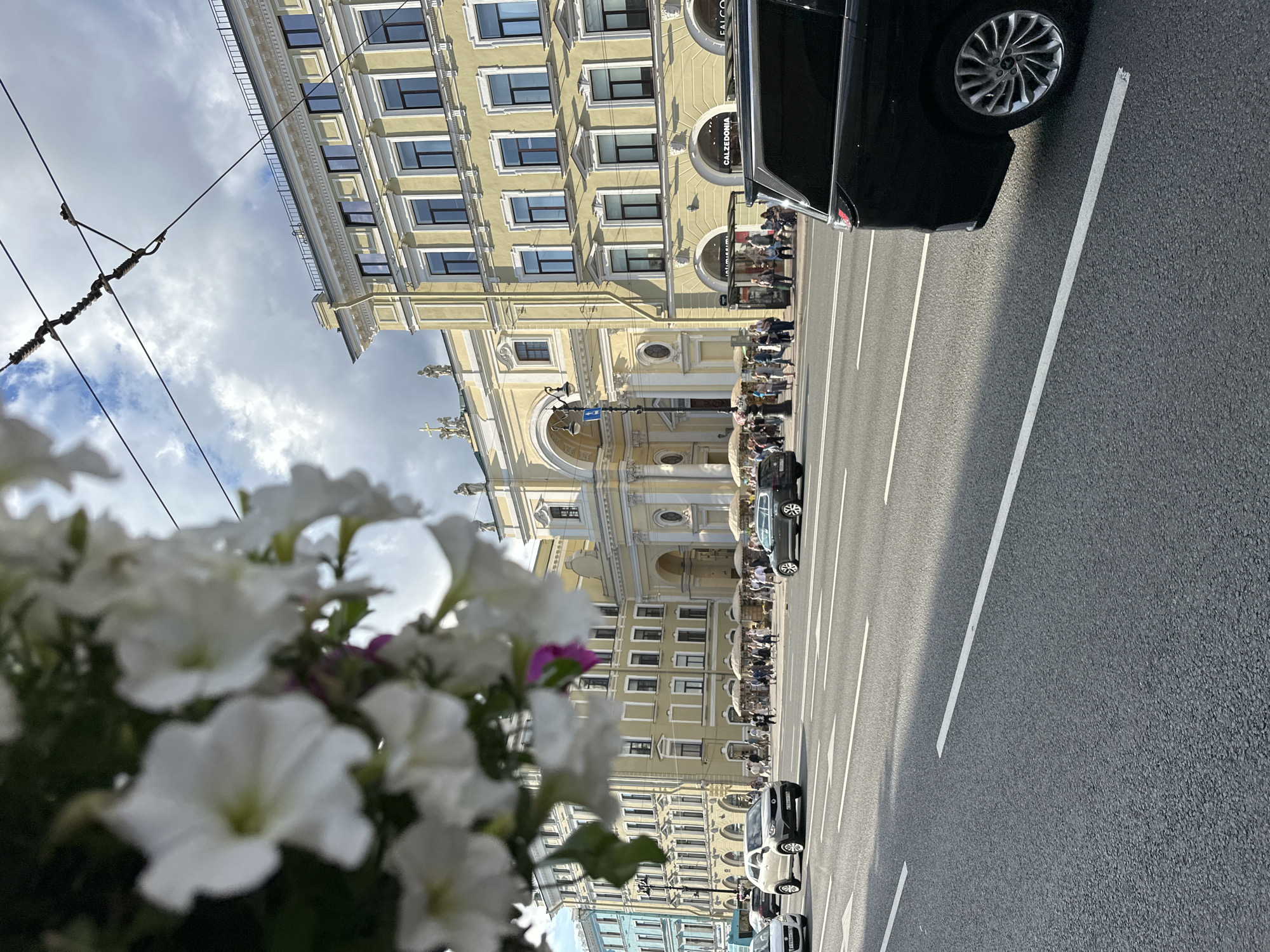 The image captures a street scene in Saint Petersburg, Russia, featuring an ornate yellow building with arched entrances and numerous windows. In the foreground, a cluster of white petunias is blurred, creating a soft, natural frame for the urban landscape. The building's architecture suggests a historical or classical style, with decorative elements along its facade.
Several cars are visible on the asphalt road, which has distinct white lane markings. One black car is prominently parked in the upper right portion of the frame, with its wheel and part of its body visible. Further down the street, more vehicles are lined up, some appearing to be moving or stopped in traffic.
On the sidewalk in front of the building, a number of people are gathered, suggesting a public space or a gathering. Some are walking, while others appear to be congregating near what looks like storefronts or cafes. The presence of umbrellas suggests a possibly sunny but potentially changeable day, or perhaps outdoor seating arrangements.
The sky is partly cloudy, with patches of blue sky visible amongst the white and gray clouds, indicating daytime. The lighting suggests it is likely midday or afternoon, with shadows cast by the building and vehicles. The overall mood is one of a bustling urban environment, with a blend of historical architecture and modern city life. Some text is visible on awnings of shops in the building, including "CALZEDONIA" and "FALCONERI".