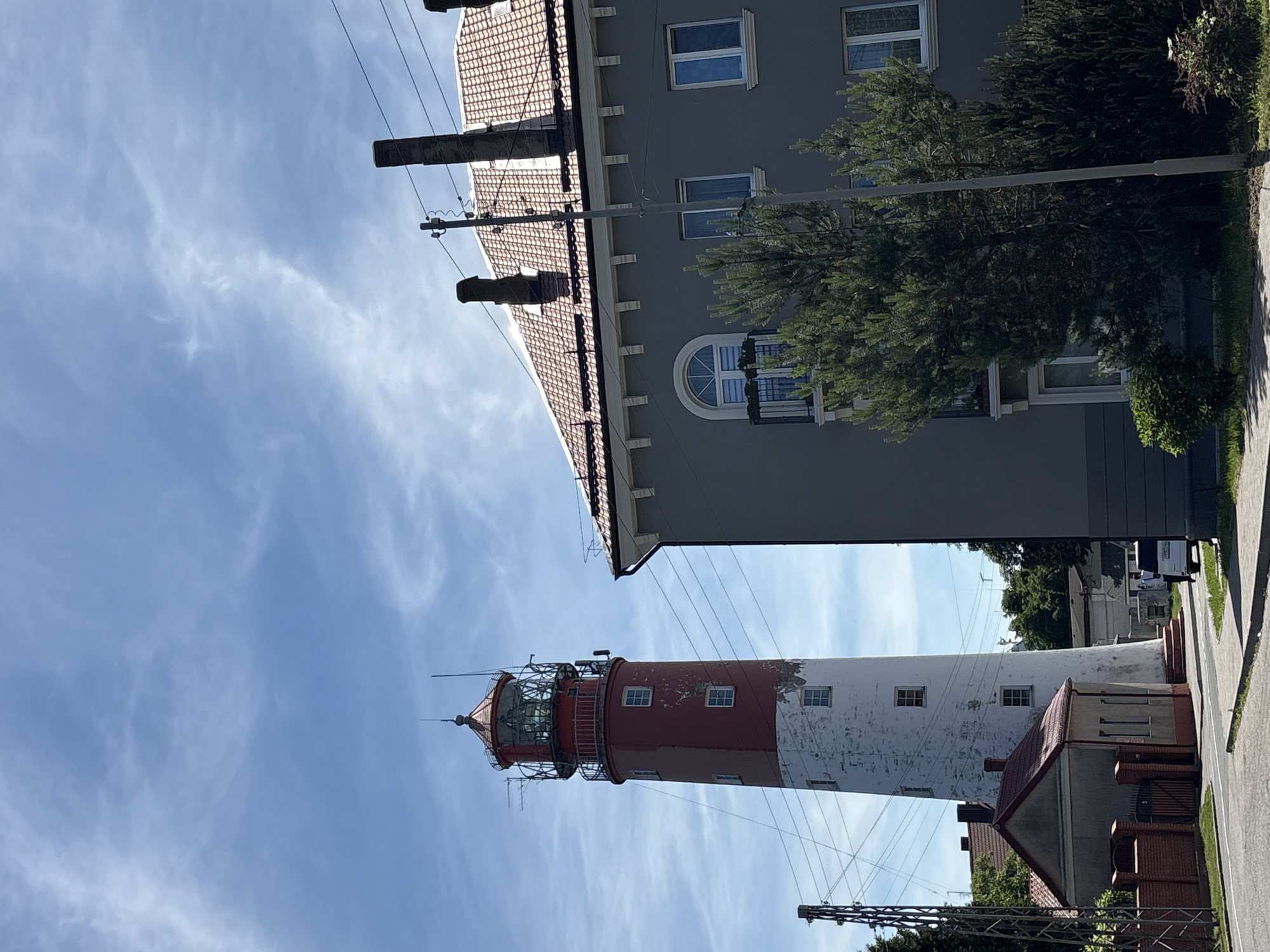 The image shows a lighthouse in Baltiysk, Russia, a port city located on the Baltic Sea. The lighthouse is a tall, cylindrical structure, painted white and red, with a lantern room at the top. It stands next to a modern building with a red tiled roof and several windows. A smaller, gabled building with a red roof is situated at the base of the lighthouse. The scene is set against a backdrop of a blue sky with scattered white clouds. Power lines and a street light are visible in the foreground, and a white van is parked on the side of the road. The vegetation includes trees and bushes. The overall impression is one of a coastal town with a mix of historic and contemporary architecture.