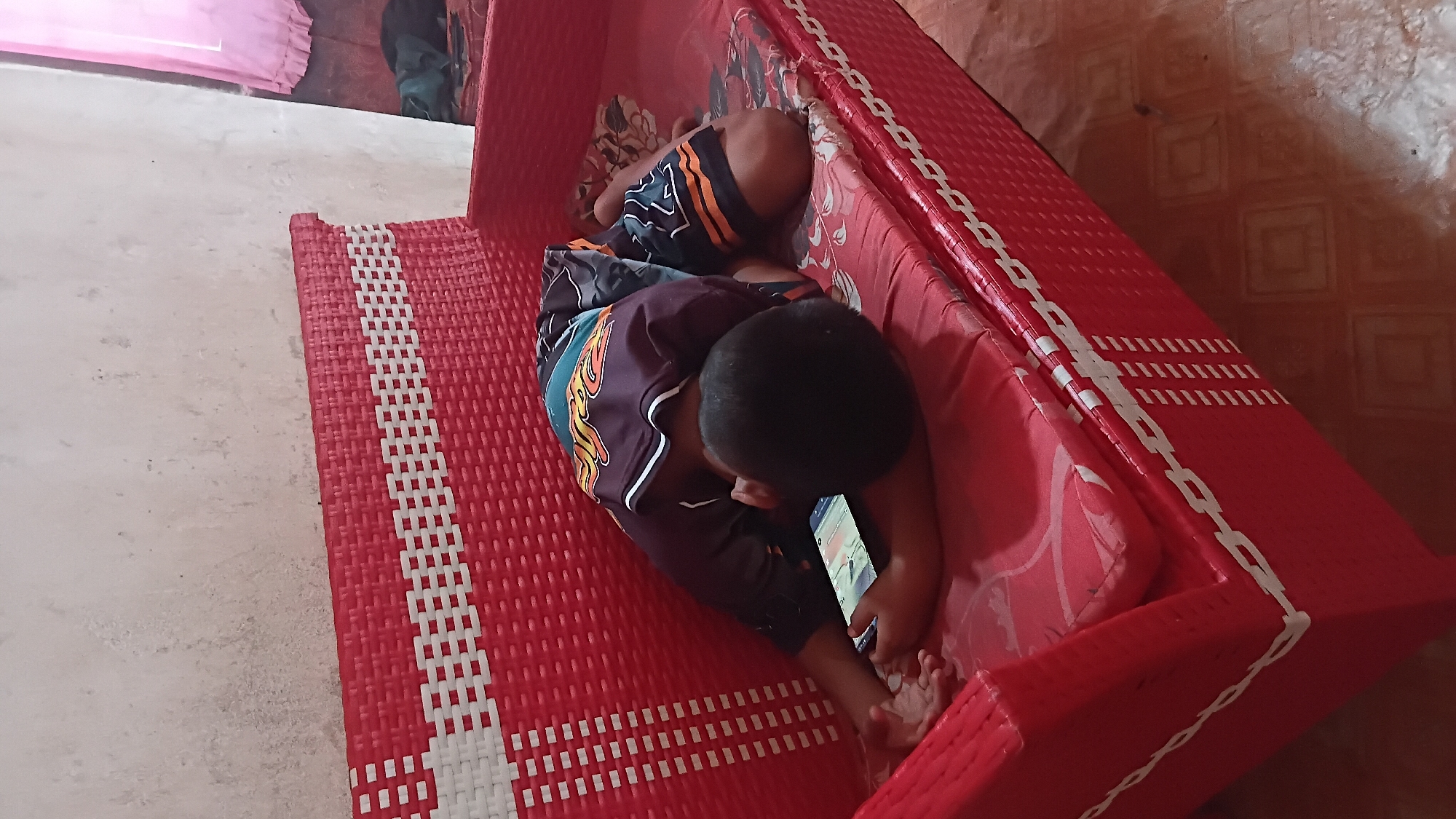 A child is lying face down on a red woven bench, engrossed in a mobile phone. The child is wearing a dark t-shirt with colorful graphics and dark shorts with an orange and white stripe pattern. The bench is positioned against a light-colored, textured wall. A pink curtain is visible in the upper right corner of the image. The floor appears to be tiled. This scene likely depicts a domestic setting within a home in Prabumulih, Indonesia.
