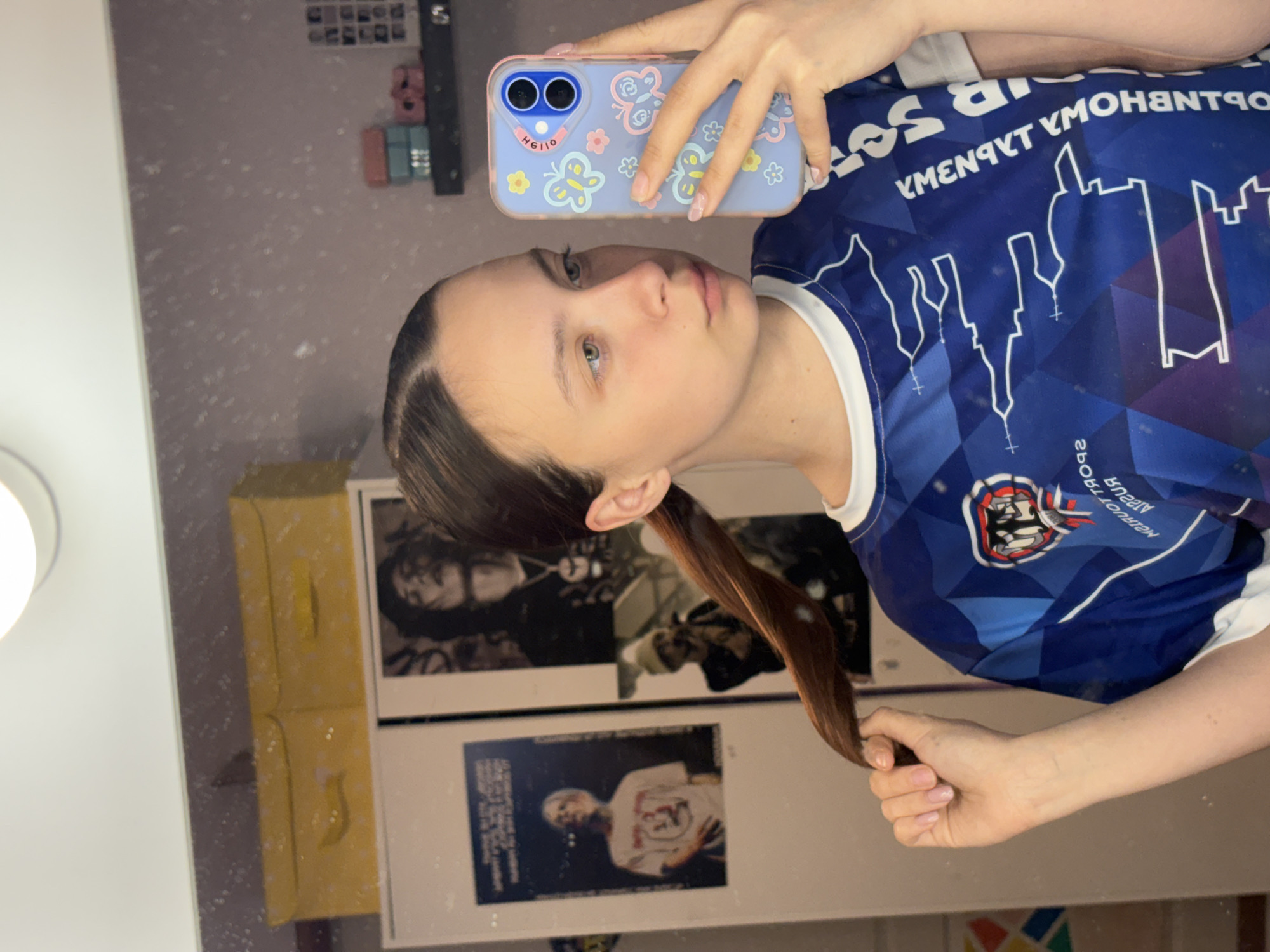 A young woman is taking a selfie in front of a mirror. She is holding a blue iPhone in her right hand with a light blue phone case decorated with butterflies and flowers. The woman is wearing a blue t-shirt with white trim and a blue and white graphic design that appears to depict the skyline of a city, potentially Прикамский, Russia, given the text on the shirt. Her hair is partially pulled back into a ponytail. Behind her, there are yellow boxes and framed posters of men.