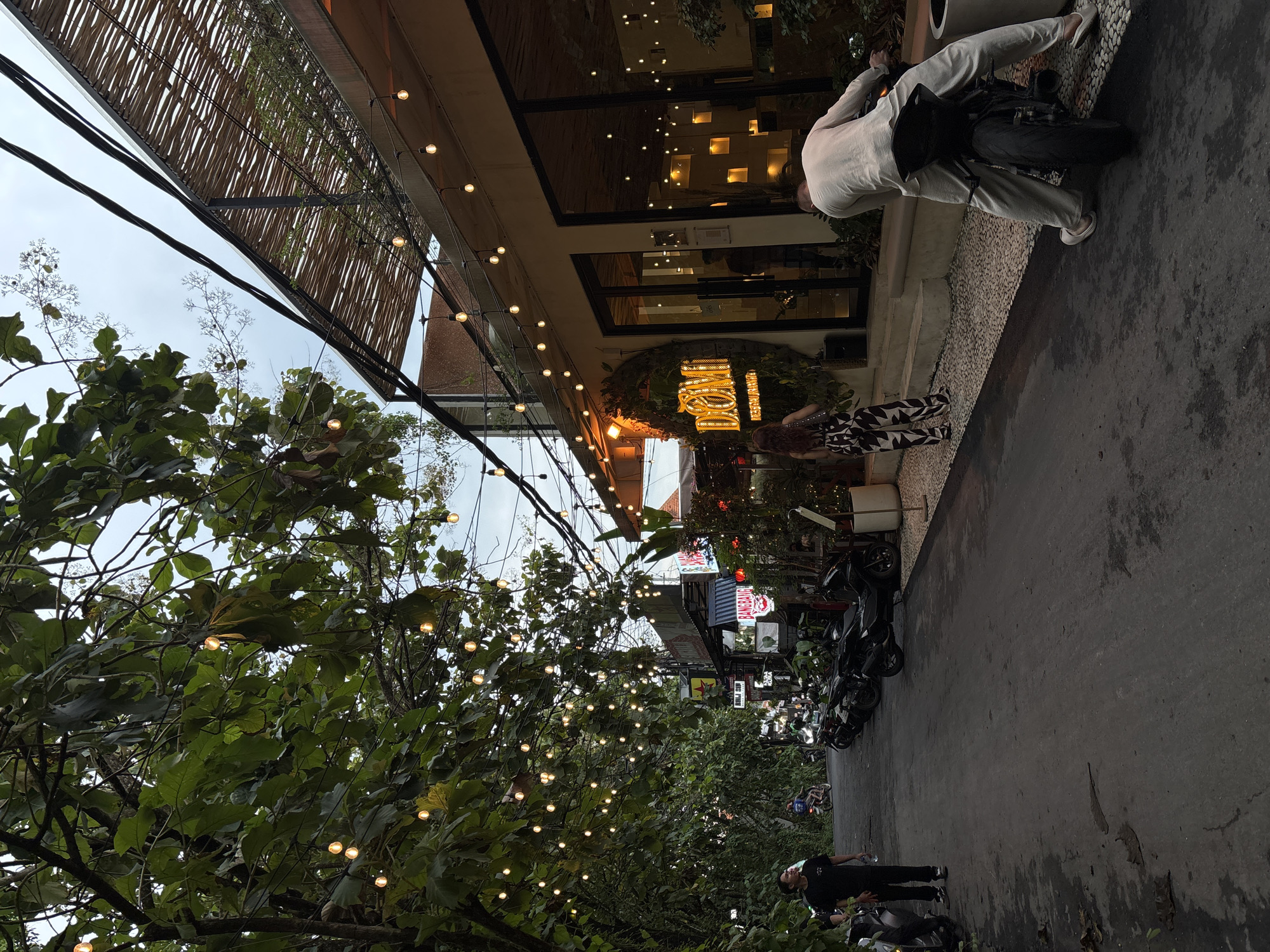 The image captures a vibrant street scene at dusk or early evening, likely in North Kuta, Indonesia. Lush green trees line the left side of the street, adorned with numerous warm-toned string lights that create a festive ambiance. Overhead, power lines crisscross against an overcast sky.
On the right, a modern building, possibly a restaurant or cafe, features a large glass facade reflecting interior lights. A bamboo-like canopy extends from the building. Below this canopy, a prominent illuminated sign displaying the text "BUMBU" in a decorative font is visible, framed by greenery.
Several people are present in the scene. One individual in light-colored attire (white shirt, light pants) is leaning over a black motorbike parked on the sidewalk, appearing to be engaged with it. Nearby, another person with long, dark, wavy hair stands near the building's entrance, wearing a black and white patterned outfit. Further down the street, a person in a dark t-shirt and pants stands near another motorbike. A row of parked motorbikes lines the street in front of the businesses.
The overall atmosphere is calm and inviting, with the artificial lights providing illumination as natural light fades. The presence of motorbikes and the architectural style are strong indicators of the Indonesian setting. Other commercial signs are visible further down the street, with "KINGBANG" partially legible on one.