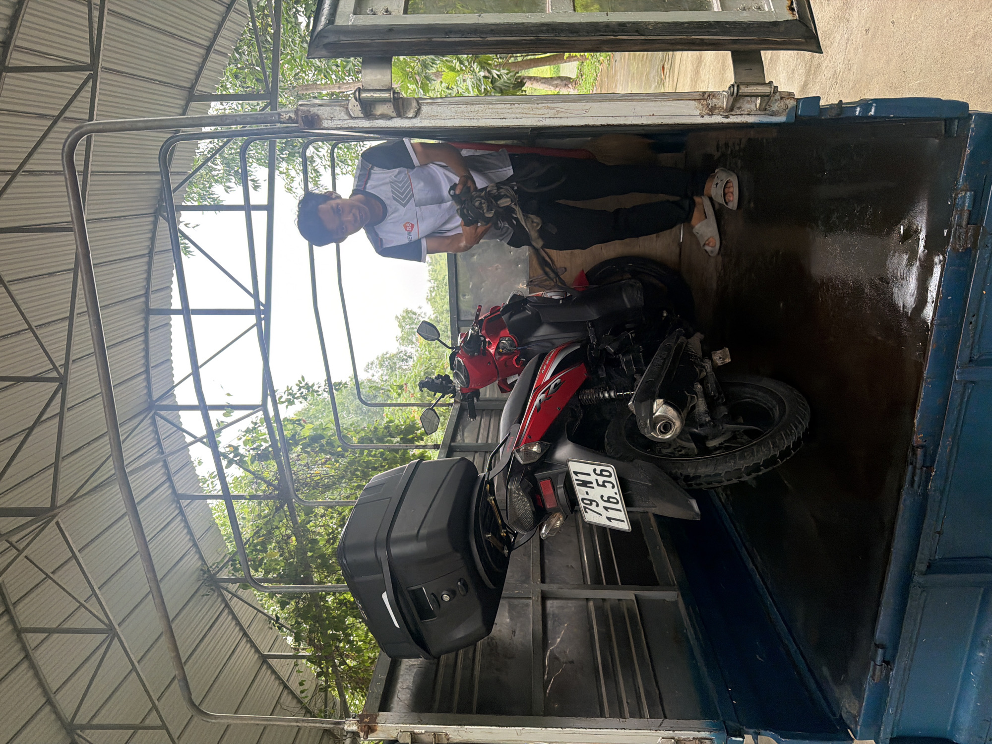 In the city of Khanh Vinh, Vietnam, a young man stands inside a large vehicle, possibly a truck or van, with a metal roof structure. He is holding a dark, cloth-like object in his hands, appearing to be cleaning or wiping something. To his left, a red motorcycle with a black storage box is parked, its license plate reading "79-N1 116.56". The floor of the vehicle appears wet. The overall environment suggests an outdoor setting, with greenery visible in the background.