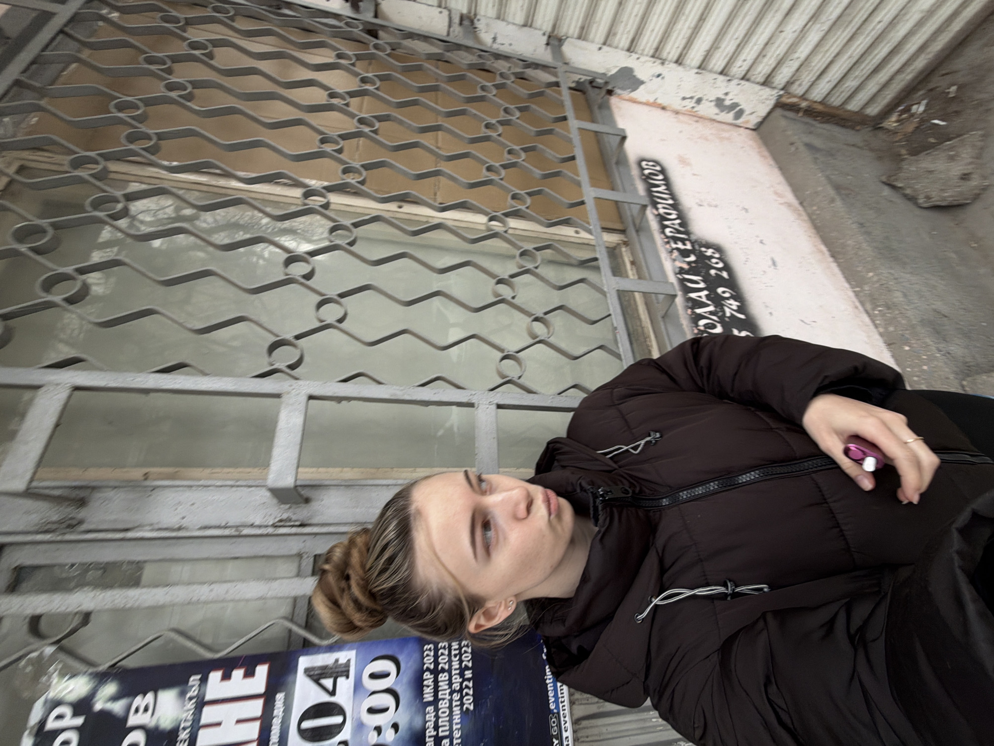 The image shows a woman in a dark brown puffer jacket, with her hair in a bun, standing outside a building in Plovdiv, Bulgaria. The building has a metal grate over the window and a sign with the name "Олай Серафимов" and the phone number "5 749 268". There is also a poster visible with the words "ДНИ НА МЕДИЯ," "ЕКТАКЪЛ," "HE," "04:00," "ИКАР 2023," "ПЛОВДИВ 2023," "ТЕТНИТЕ АРТИСТИ 2022 и 2023," and "eventim". The woman is holding a small pink object in her hand.