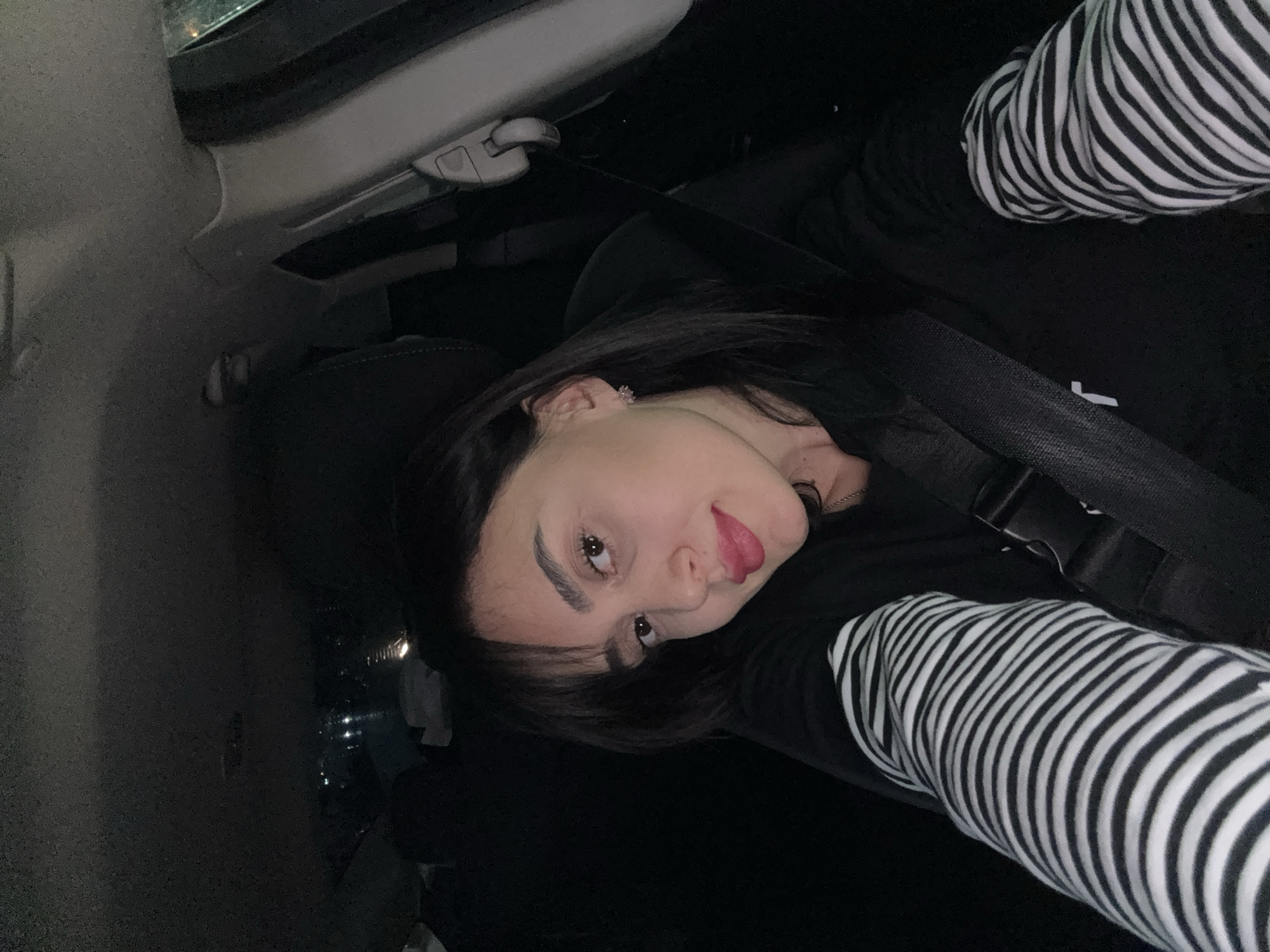 A young woman is positioned in the passenger seat of a car. She is wearing a seatbelt. She has dark hair and is wearing a long-sleeved black shirt under a black and white striped shirt. The interior of the car is dark. It is not possible to determine the location in the city of Tbilisi, Georgia, from the image.