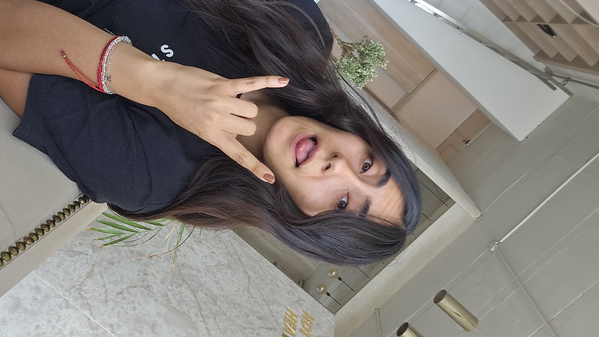 The media file displays a portrait-oriented image rotated 90 degrees clockwise, featuring a young woman posing playfully indoors. She has long, dark hair and is looking directly at the camera with a mischievous expression, sticking out her tongue. Her right hand is raised, forming a "rock on" hand gesture, and her fingernails are painted a light brown color. On her left wrist, she wears layered bracelets: one thin red string bracelet and two silver-colored beaded or chained bracelets. She is wearing a black short-sleeved top.
The background suggests a stylish modern interior, likely located in Puebla, Mexico. The floor is covered with light-colored, possibly marble-patterned tiles. A light-toned piece of furniture, perhaps a sofa or armchair, with decorative brass studs along its edge, is visible. A green palm frond is seen near the floor. In the upper background, there are light-colored walls and a partial view of a wooden slatted ceiling. Two modern, gold-colored cylindrical light fixtures hang from the ceiling. Gold-colored three-dimensional letters spelling "HOT HEAVEN" are affixed to a wall. A small green plant with tiny white flowers is also visible in the upper portion of the frame. The lighting appears bright and even, contributing to a cheerful and casual atmosphere.