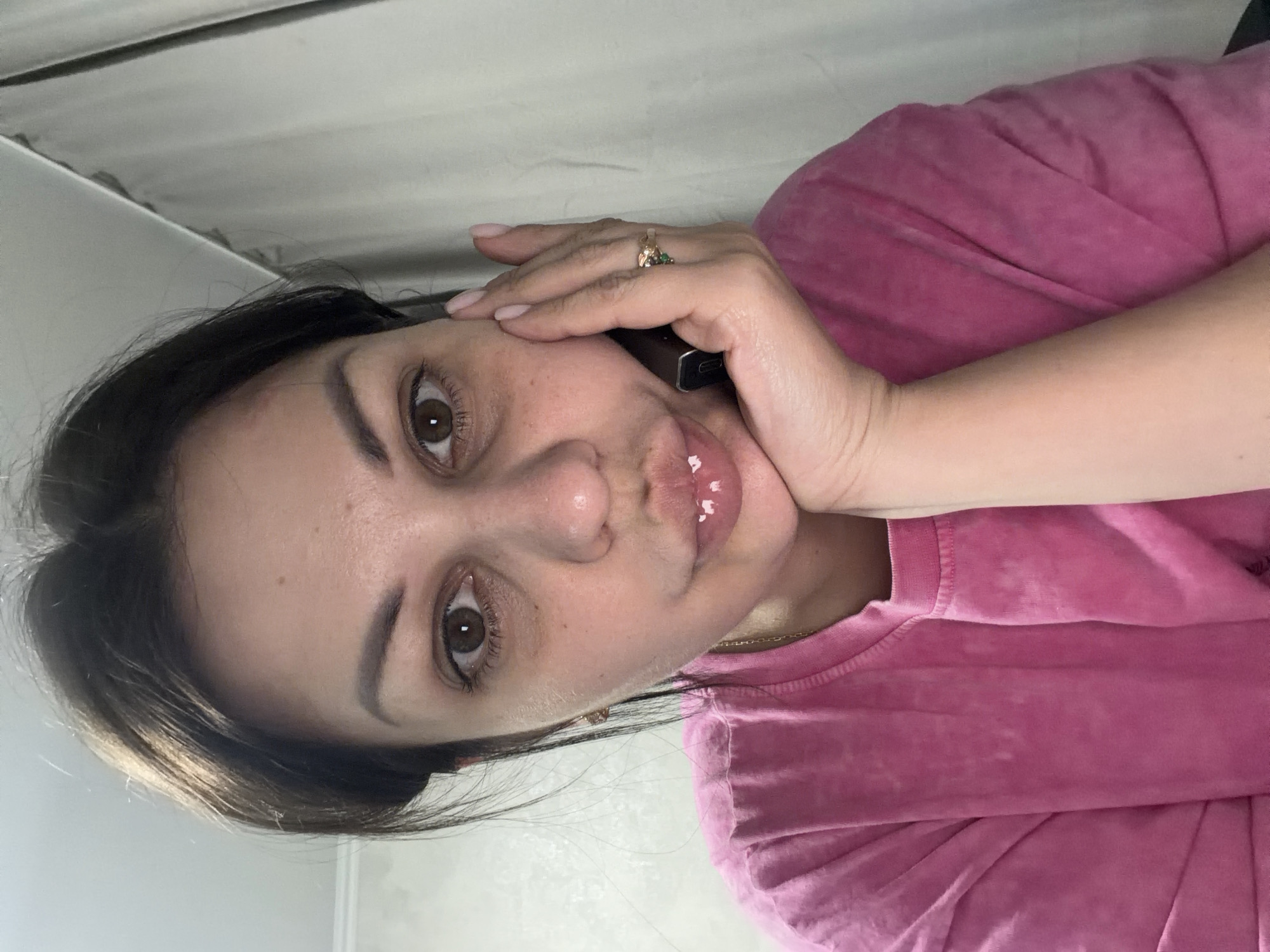 A woman is captured in a close-up selfie. She has dark hair pulled back, revealing her face. Her right hand is raised to her cheek, and she is holding a dark object, possibly a phone, to her ear. She is wearing a bright pink t-shirt. Her lips are slightly parted, and a few white specks are visible on them. She has brown eyes and appears to be looking directly at the camera. The background is indistinct, with a textured wall and what appears to be a curtain or drape. There is no discernible connection to Sochi, Russia, in this image.