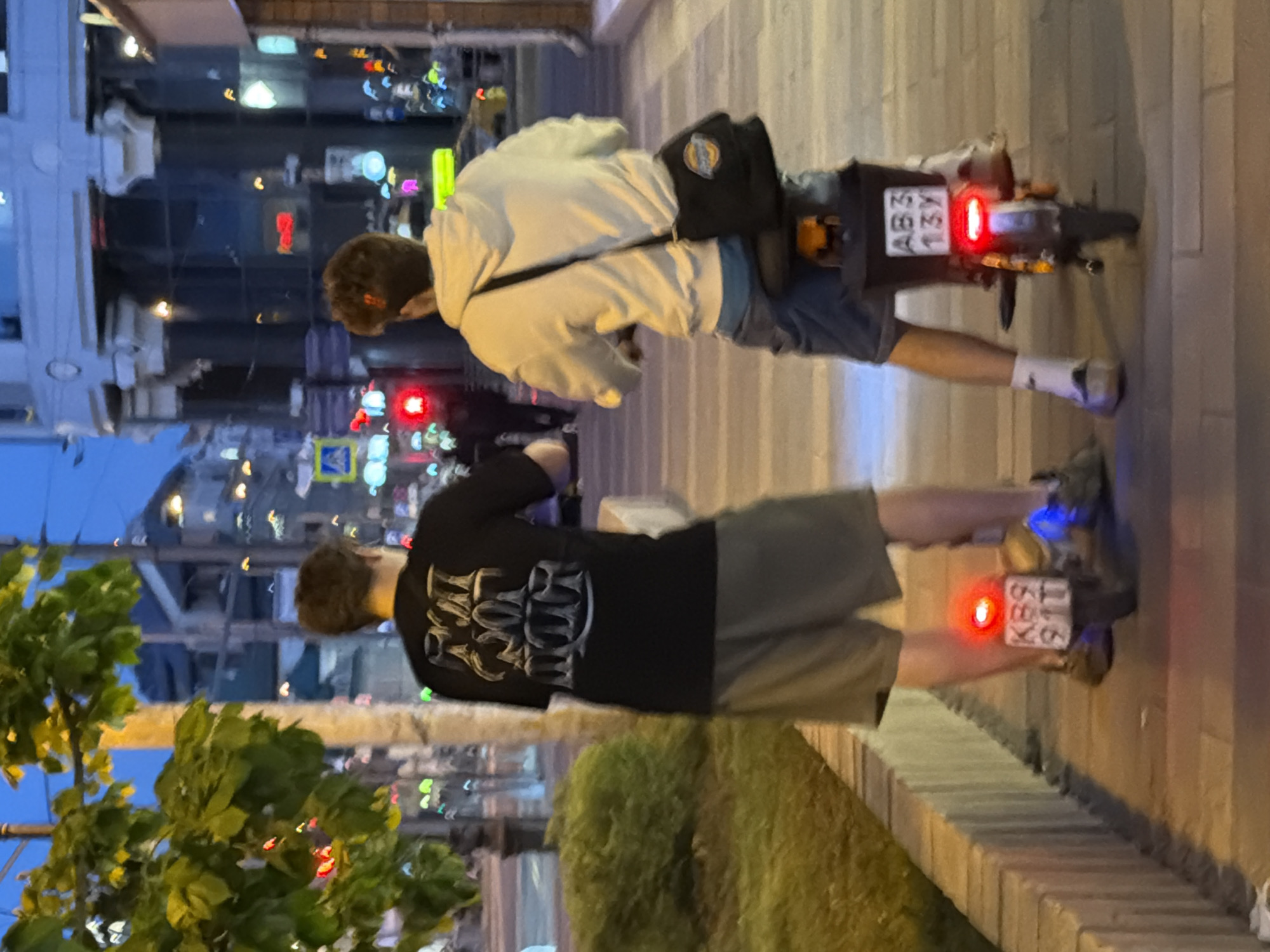 Two people ride electric scooters down a sidewalk in Moscow, Russia. The people are male, and dressed casually. One wears a white sweatshirt and the other a black shirt. Both wear shorts and sneakers. The electric scooters have red taillights. The license plates are visible and read “A 131 BA 799” and “K892 TT 799”. The buildings in the background show commercial signage and apartment structures. The street lights are on. Bushes are along the sidewalk.