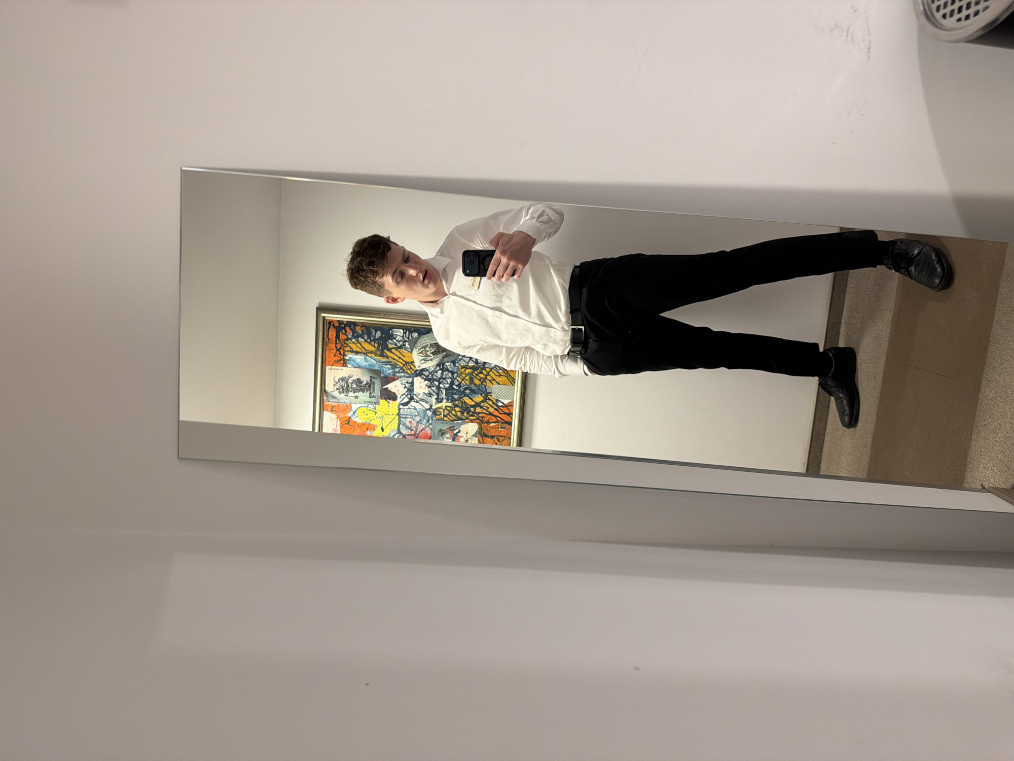 A young man stands in front of a mirror, taking a picture of himself with his smartphone. He is wearing a white dress shirt, black pants, and black shoes. Behind him is a framed, colorful, modern abstract painting. The setting appears to be an indoor space, likely a bathroom or changing room. The exact location within Varna, Bulgaria, cannot be determined from the image.