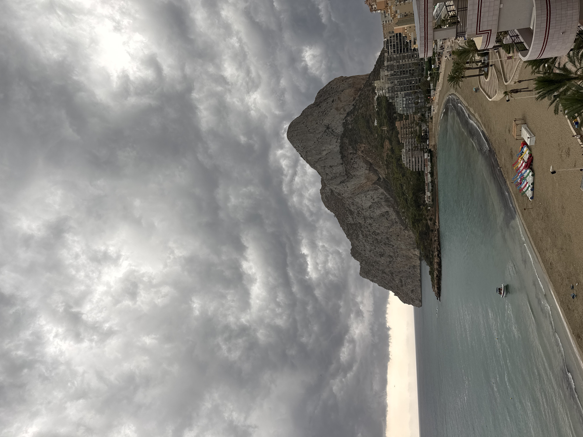 The image depicts a coastal scene in Calp, Spain, dominated by the imposing Peñón de Ifach rock formation. The sky is overcast with heavy, dark gray clouds, creating a dramatic atmosphere. A calm, turquoise sea laps against a sandy beach lined with colorful paddle boats. Several buildings, including apartment complexes and a hotel with balconies, are visible along the coastline, nestled at the base of the rock. A small boat with people on board is anchored in the shallow water. The beach appears sparsely populated, with a few individuals walking or lounging. The foreground shows a section of the beach with palm trees and part of a building's facade. The overall impression is one of a somewhat moody, overcast day at a popular coastal resort.