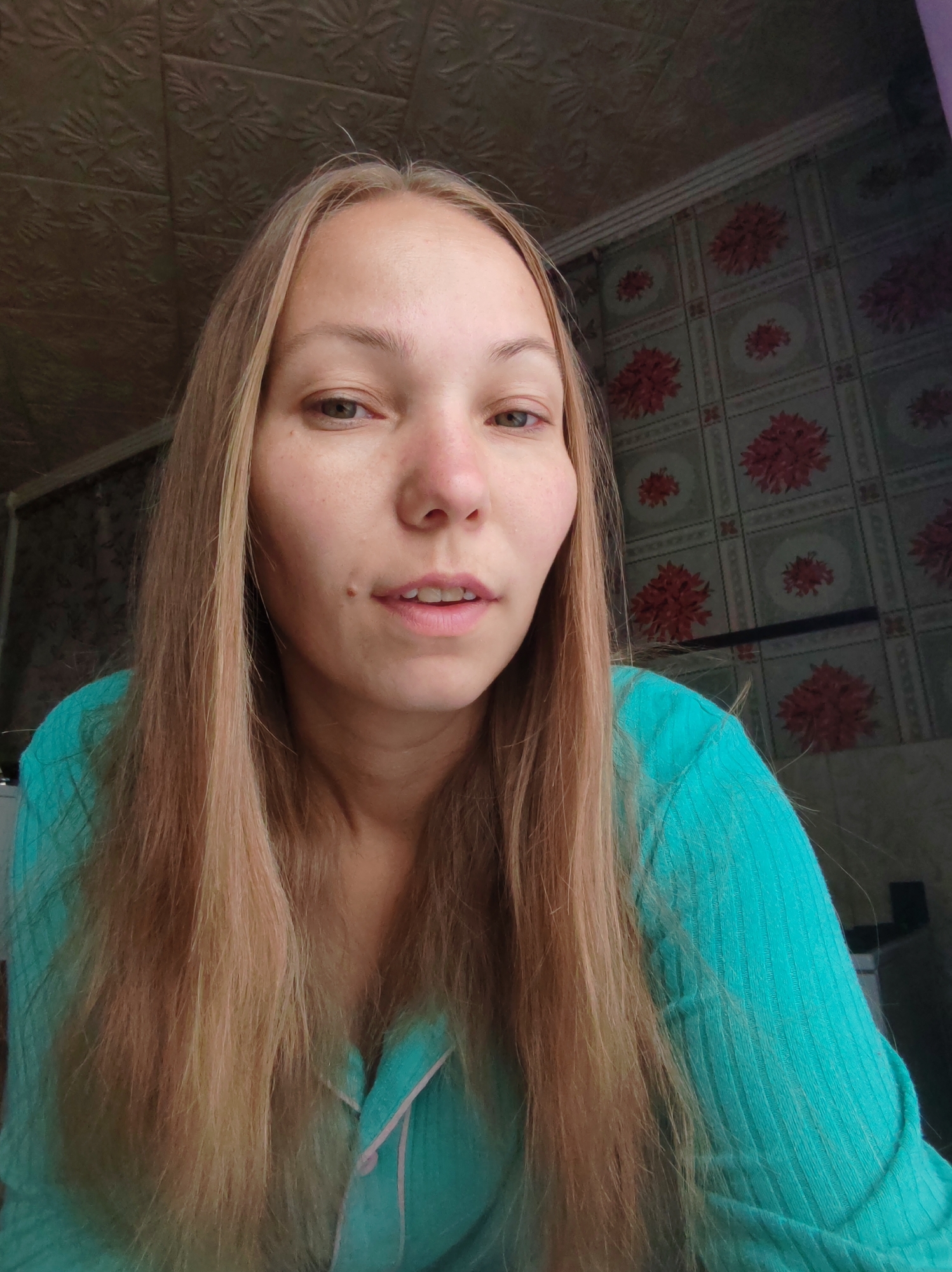 The image shows a close-up, waist-up shot of a young Caucasian woman with long, blonde hair. She is wearing a turquoise, ribbed shirt with pink trim. Her head is slightly tilted, and she is looking directly at the camera. Her expression is neutral, with her mouth slightly open. The background consists of a patterned wall and a textured ceiling, suggesting an indoor setting. There is no indication of the specific location within the city of Chuchlem, Russia.