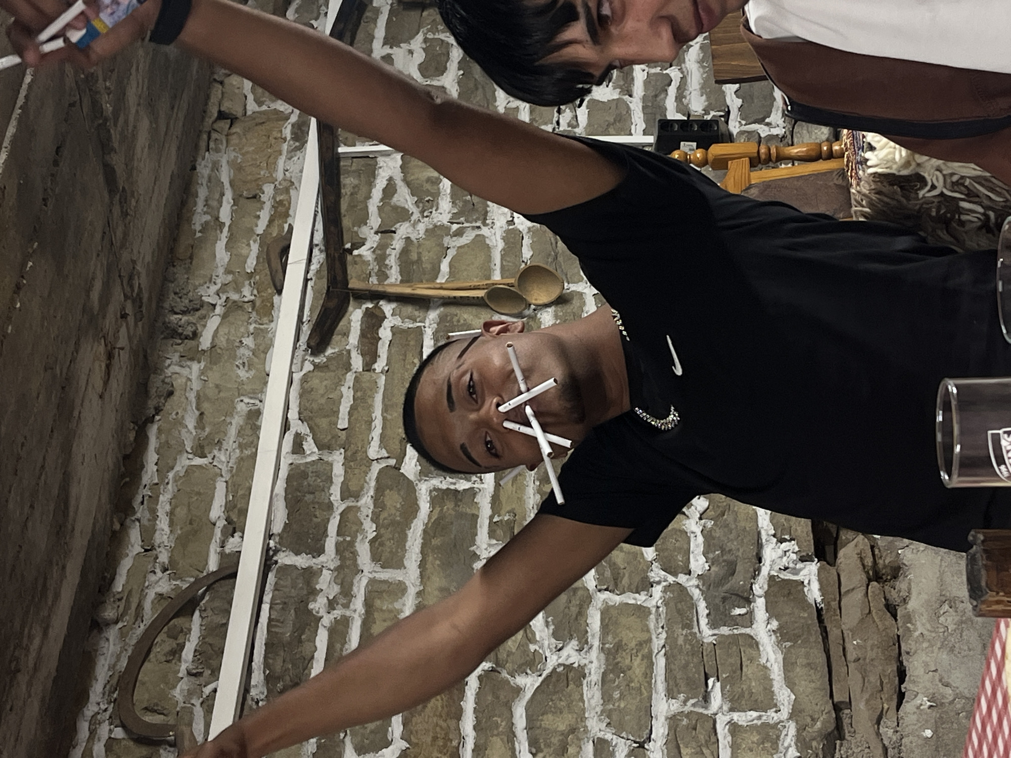The image shows two young men in what appears to be an interior space with stone walls. The main subject, a young man in a black t-shirt with a Nike swoosh, has several cigarettes stuck in his mouth and nose. He's holding up what appears to be a cigarette pack. Another young man is partially visible at the top right of the frame. The background includes various rustic objects hung on the stone wall, such as wooden spoons. The location is Lozarevo, Bulgaria.