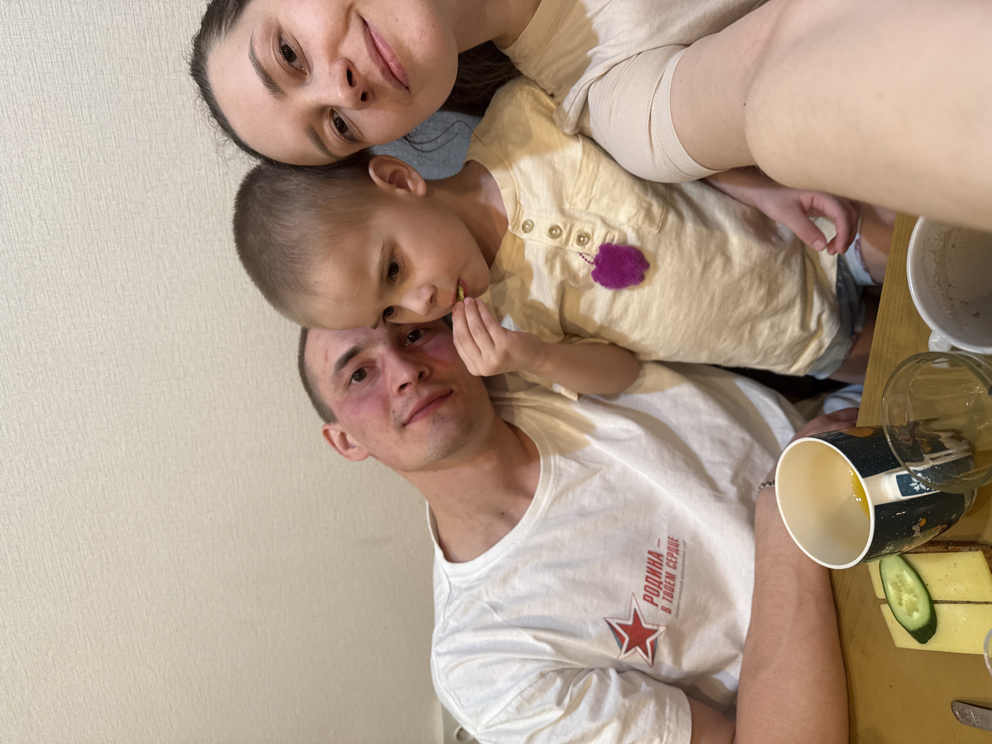 A selfie shows three people, likely a family, posing indoors. A woman is at the top, with a young boy nestled between her and a man. The boy is eating something. The man is wearing a white t-shirt with a red star and Cyrillic text, possibly indicating a patriotic or Soviet-era theme. The setting appears to be a home, with a table visible on the right side. On the table, there is a cup, a glass, bread, cheese, and a slice of cucumber. There is no information that allows determining that the location is Chaikovsky, Russia.