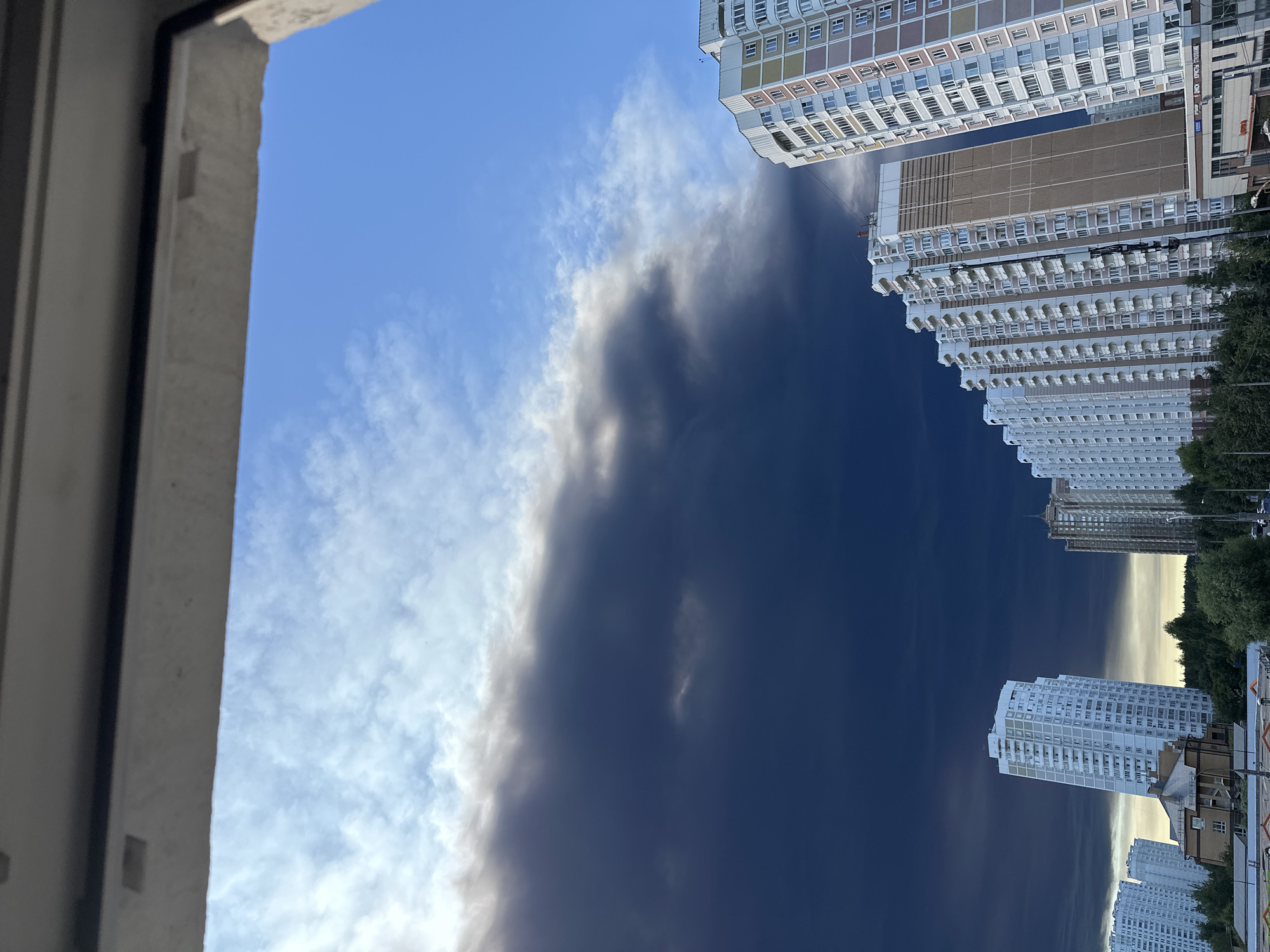 The image, taken from a window frame overlooking a city, depicts a dramatic sky filled with dark, ominous clouds that take up the majority of the frame. To the right of the image, several tall, modern apartment buildings are visible. The buildings have balconies and a mix of white and beige facades with colorful accents. The sky transitions from a bright blue at the top right to a deep, almost black hue as it moves towards the left, suggesting a storm or severe weather event. Below the sky, a line of trees separates the buildings from the darker sky. The lower part of the frame shows the tops of more buildings and some vegetation. The overall impression is one of an impending storm over a residential urban area in Podolsk, Russia.