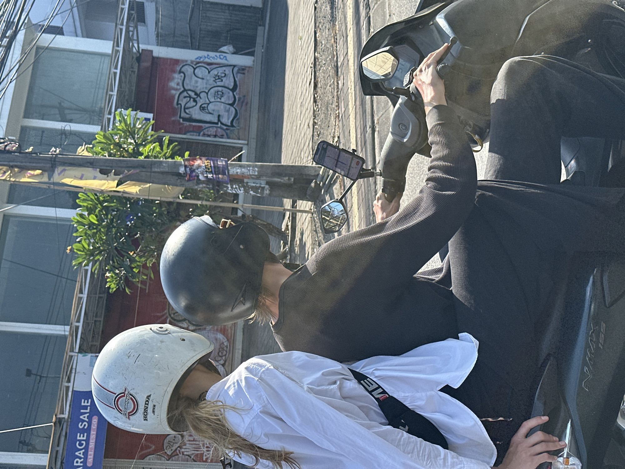 Two individuals are riding a dark-colored scooter on a street in Kuta, Indonesia. The person in the foreground, operating the scooter, wears a dark-colored long-sleeved shirt and black pants, with a black helmet. Their hands are on the scooter's handlebars, and a mobile phone is mounted on the handlebars, displaying a navigation app. The second individual, seated behind the driver, wears a white long-sleeved shirt and black pants, and a white helmet. Their left hand is resting on the driver's leg. A black strap with white lettering is visible across the white shirt. In the background, buildings with signs, including one that reads "GARAGE SALE," and graffiti are visible. A utility pole with attached wires and posters is also present. The scene appears to be a sunny day, with shadows cast on the street.