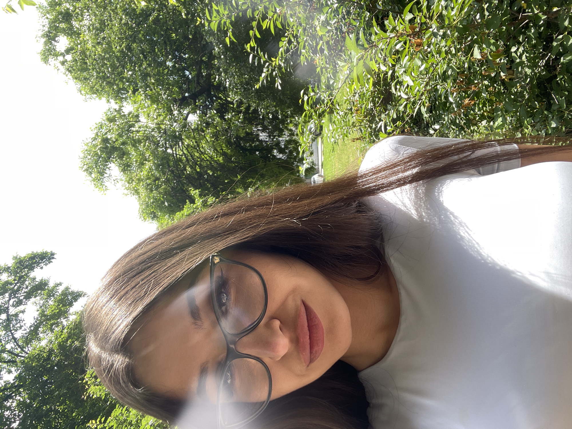 A person with long brown hair and wearing glasses is shown in a selfie. The person is wearing a light-colored shirt. The background is filled with green foliage, including trees and bushes. The location is the city of Unknown, Unknown.