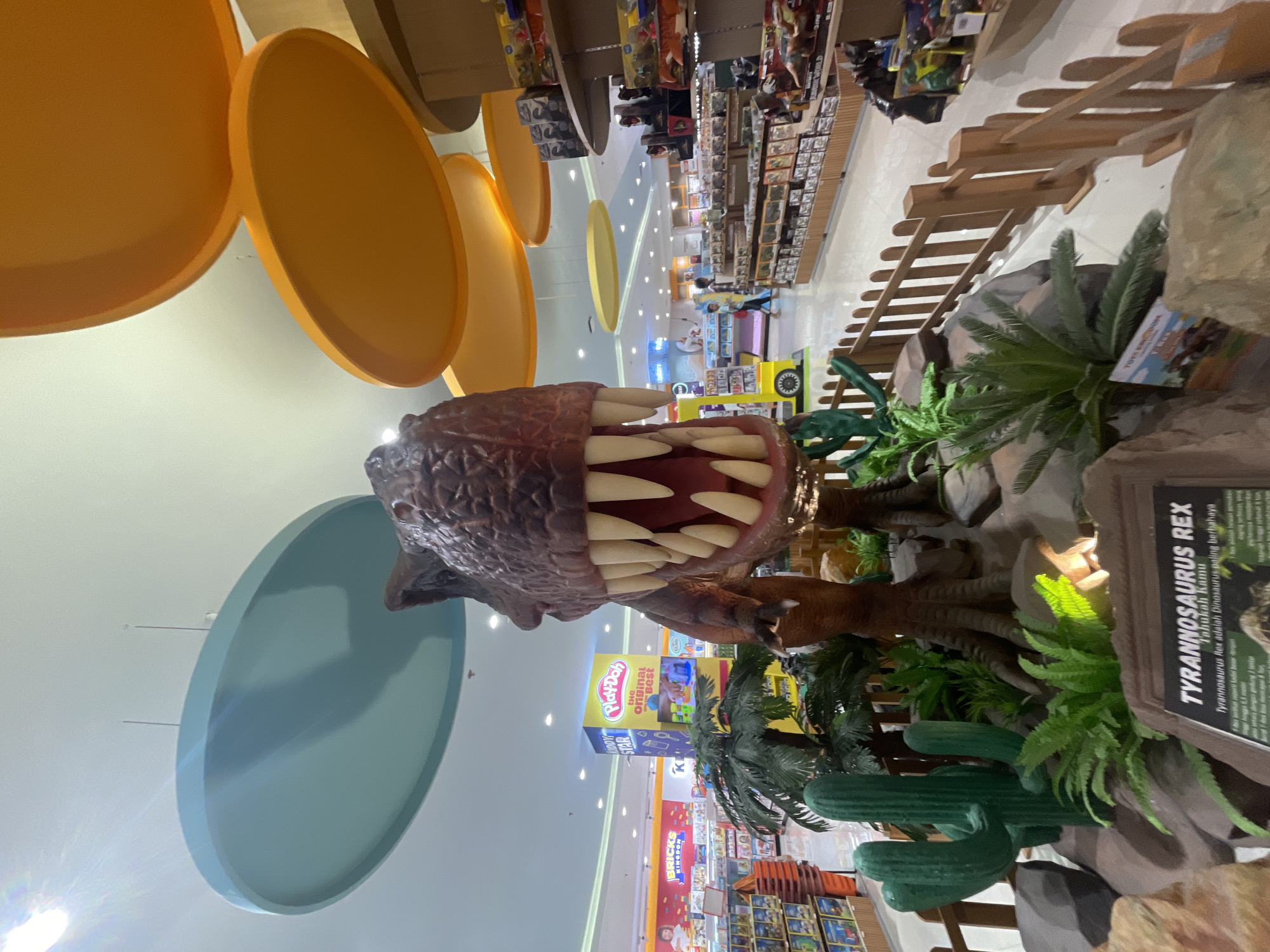The image shows a large Tyrannosaurus Rex model with open mouth inside a toy store. The model is surrounded by artificial plants and a sign stating "TYRANNOSAURUS REX Tahukah Kamu." Shelves filled with toys are visible in the background. The store appears to be located in Denpasar, Indonesia, based on the signage "Tahukah Kamu" which is in Indonesian. Large, colorful circular ceiling decorations are visible. There are signs in the background for the TOYS KINGDOM, KIDOY STAR, Play-Doh, and BRICKS. A wooden fence-like structure is in the right corner.