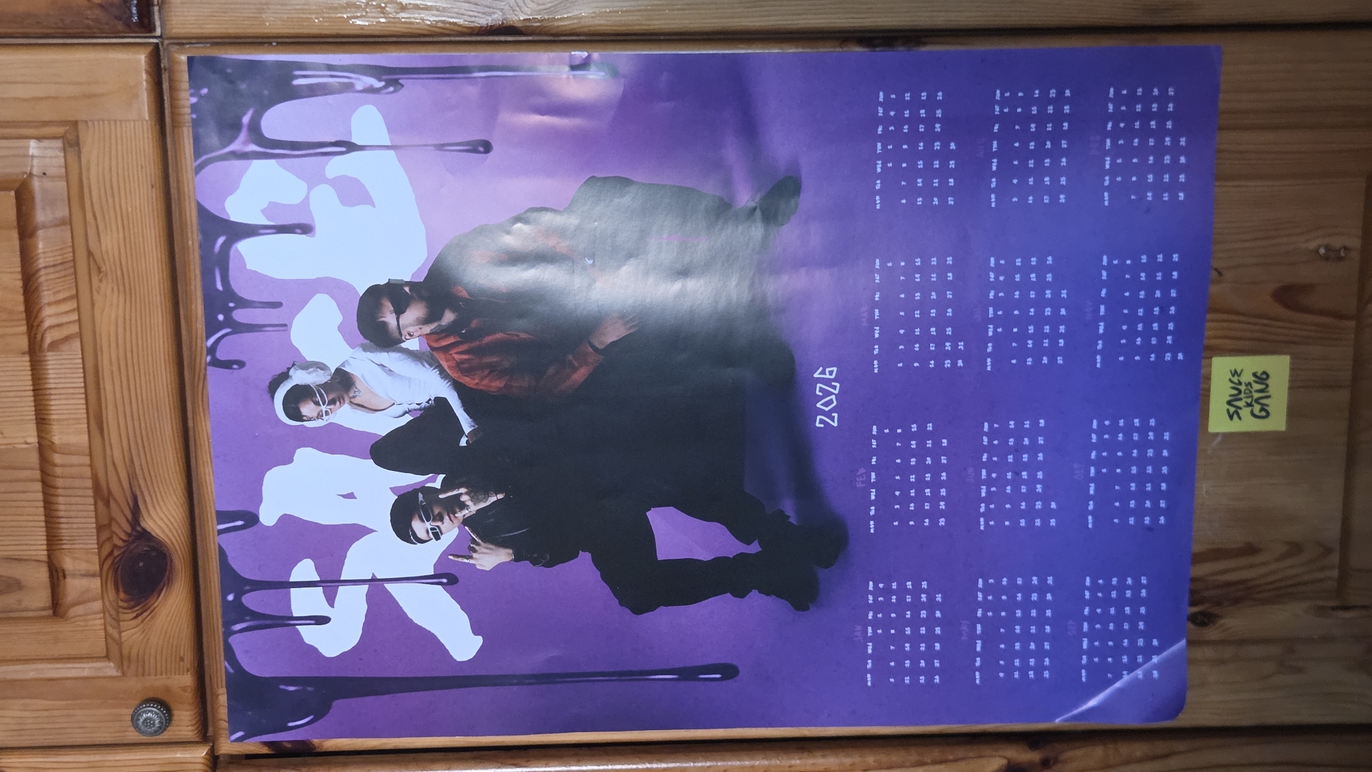 The image shows a poster of a musical group, possibly a calendar, hanging on a wooden cabinet door. The poster is purple with black drippy graphics at the top. Three people are featured in the poster. A woman in the center wears a white top and white headphones. A man on the left wears a black outfit and sunglasses, with his hands raised. A man on the right wears a red shirt and sunglasses. The calendar portion of the poster displays the year 2026 and the months of January through December. Below the poster, a yellow sticky note with "SAUCE KIDS GANG" written in black marker is attached to the cabinet. The cabinet appears to be made of light-colored wood with visible grain and knots. A circular metal knob is attached to the upper left door of the cabinet. There is no indication of the location being in Bania, Bulgaria.