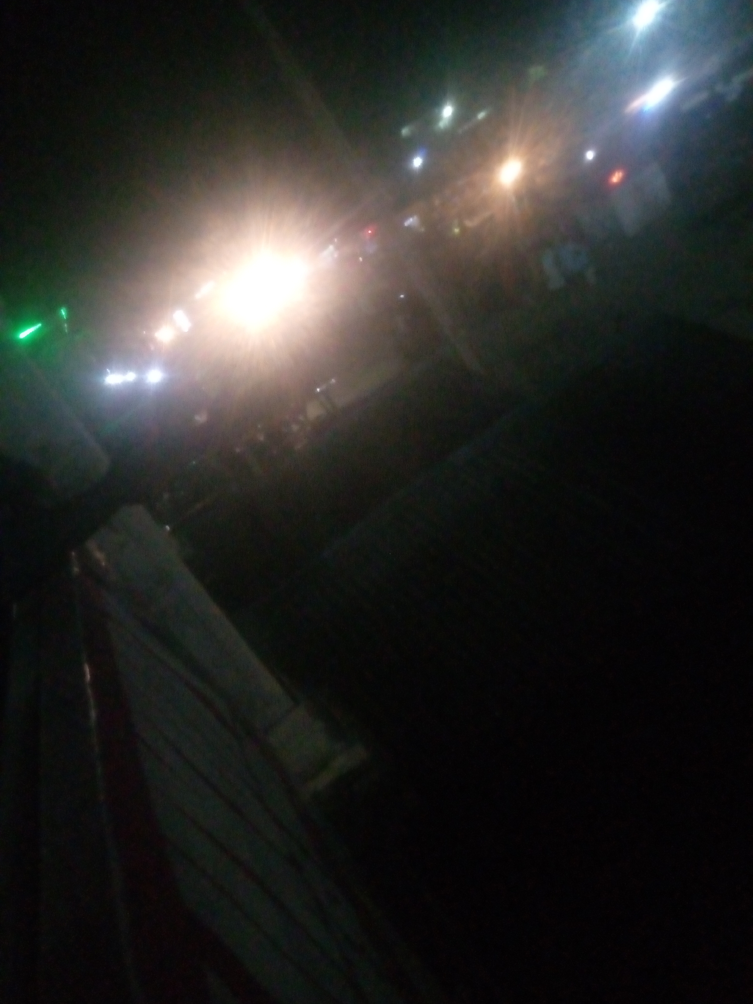 The image depicts a night scene with several bright lights, suggesting an outdoor event or a busy street in Jalingo, Nigeria. The lights are blurred and create lens flares, indicating a low-light photography situation. Some of these lights appear to be from vehicles or streetlights, with a particularly bright, yellowish light in the center of the frame. There are also some green lights visible on the left side. In the foreground, a dark, striped surface, possibly a roof or awning, occupies the bottom left portion of the image. The background is dark and indistinct, with some faint shapes that might represent people or structures. The overall impression is of a dynamic, illuminated environment at night.