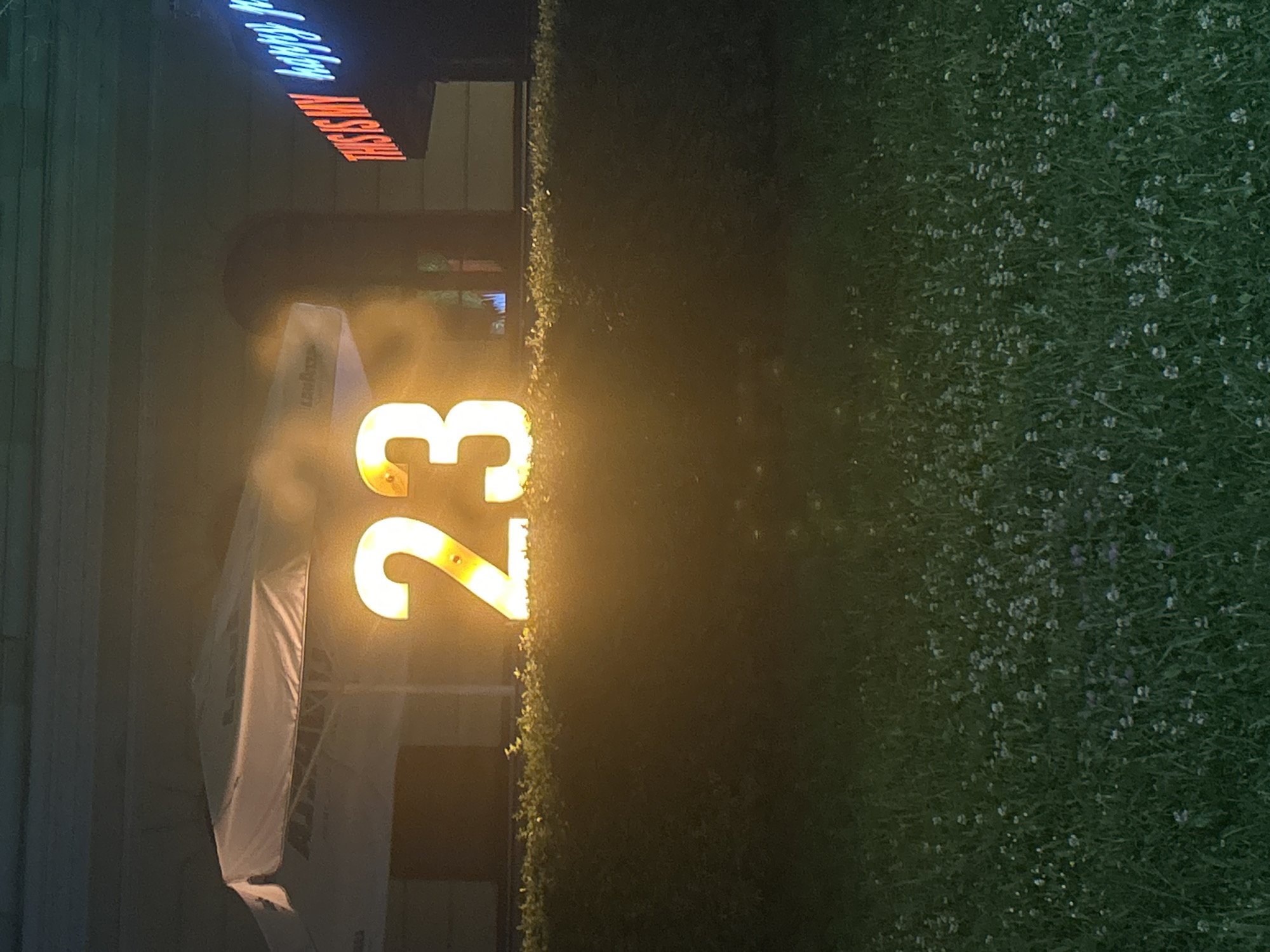 The image depicts a street scene at night. A large, brightly illuminated number "23" is visible, likely a sign or marker. Adjacent to the number is a leafy green hedge. To the left, there's a white umbrella or awning, partially obscured by fog or mist. In the background, a sign reads "THIS IS MY hobby," suggesting the location might be a business or establishment promoting a hobby. The overall scene seems to be an outdoor area in an urban environment. I cannot confirm that the location of this image is in the city of Veliko Tarnovo, Bulgaria.