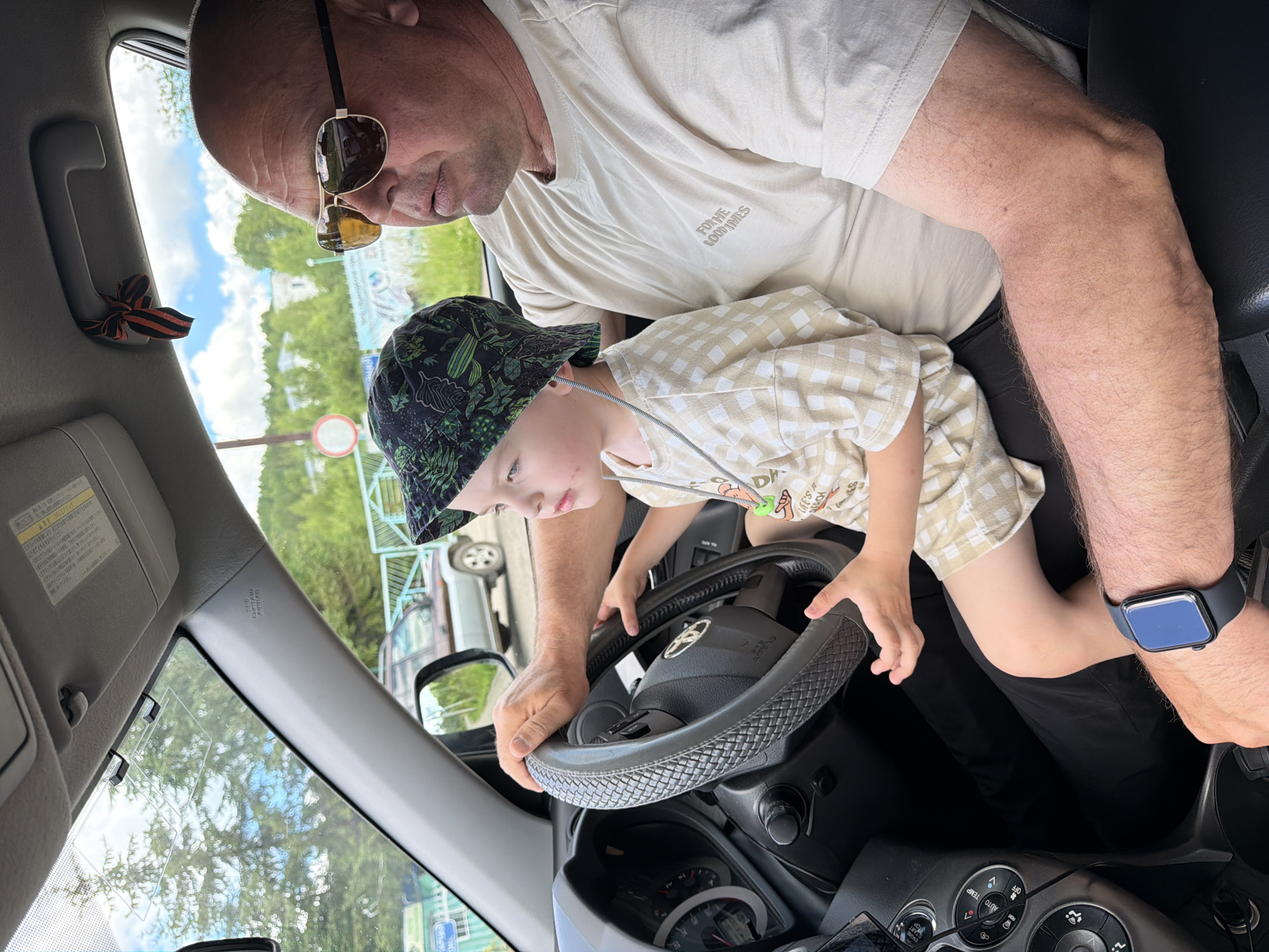 A man wearing sunglasses and a beige t-shirt sits in the driver's seat of a car, with a young child on his lap. The man's left hand rests on the steering wheel, and the child's hands are also on the wheel. The child wears a patterned bucket hat and a checkered shirt. The car's interior is visible, including the dashboard and steering wheel. Outside the car, trees and a blue sky with clouds can be seen. A decorative ribbon with orange and black stripes is tied to a handle above the windshield. The scene is likely captured in the city of Kul'dur, Russia.
