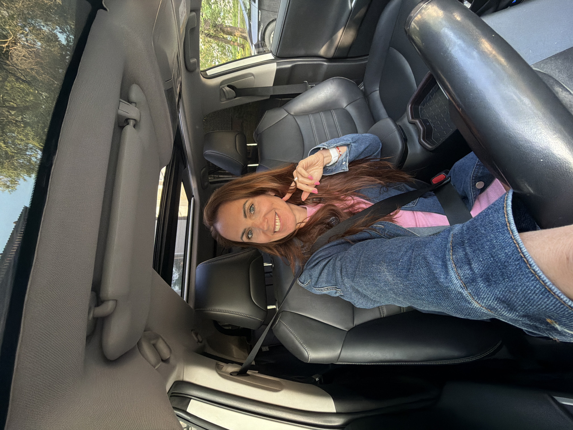 In the image, a woman with long, auburn hair and a bright smile is seated in the driver's seat of a car. She is wearing a denim jacket over a pink shirt. The interior of the car is visible, with black leather seats and a sunroof. The woman is looking at the camera and has her left hand near her face, touching her cheek. Her seatbelt is fastened. The car appears to be parked outdoors, with trees and another car visible through the windows, suggesting an urban or suburban environment. The provided crops confirm the interior details of the car, including the sunroof, visors, leather seats, and seatbelt. The scene does not provide any specific indicators placing it in Birobidzhan, Russia.