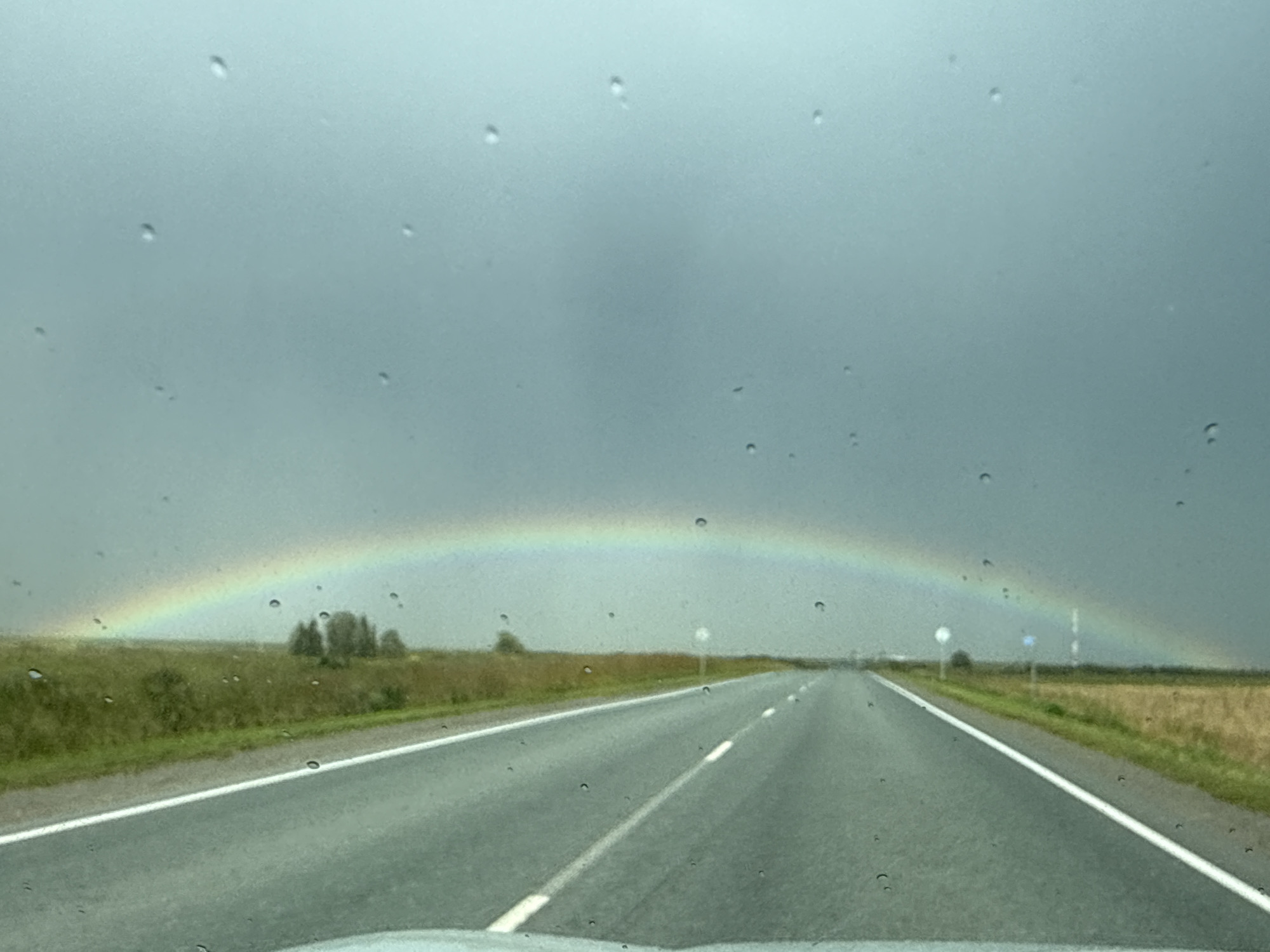 The image shows a view from a car driving on a road through a rural landscape under a cloudy sky with a rainbow. Raindrops are visible on the windshield, distorting the view. The road is paved and has white lines marking the lanes. On either side of the road are fields of green grass and dry vegetation. In the distance, a cluster of trees is visible, and a faint rainbow arcs across the sky. There are no people or animals visible in the image. The scene suggests a recent or ongoing rain shower. The location is identified as the city of Chuvarina, Russia.