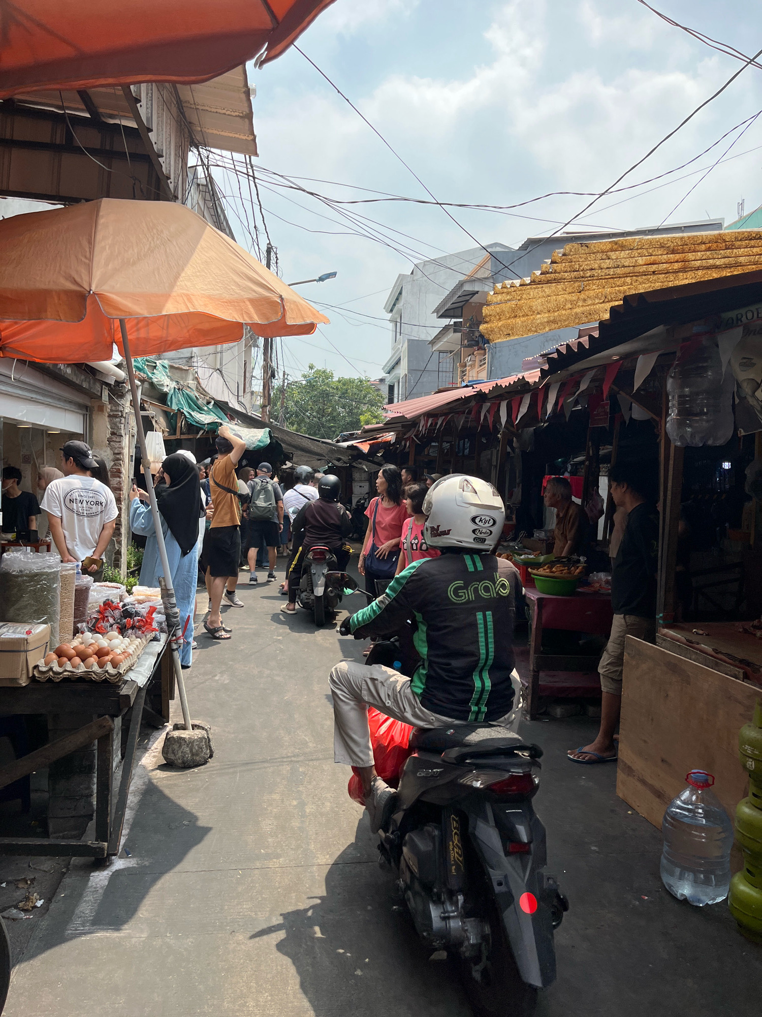 A busy outdoor market scene unfolds under a partly cloudy sky. The foreground is dominated by a person on a motorcycle, viewed from behind, wearing a white helmet with a green and black jacket that has "Grab" logo. To the left, a market stall displays eggs and other goods under an orange umbrella. Further down the narrow street, other vendors and shoppers are visible. Shops with awnings and corrugated metal roofs line the sides of the street, with various goods and people interacting. Electrical wires crisscross overhead. The ground is paved, and shadows indicate a sunny day.